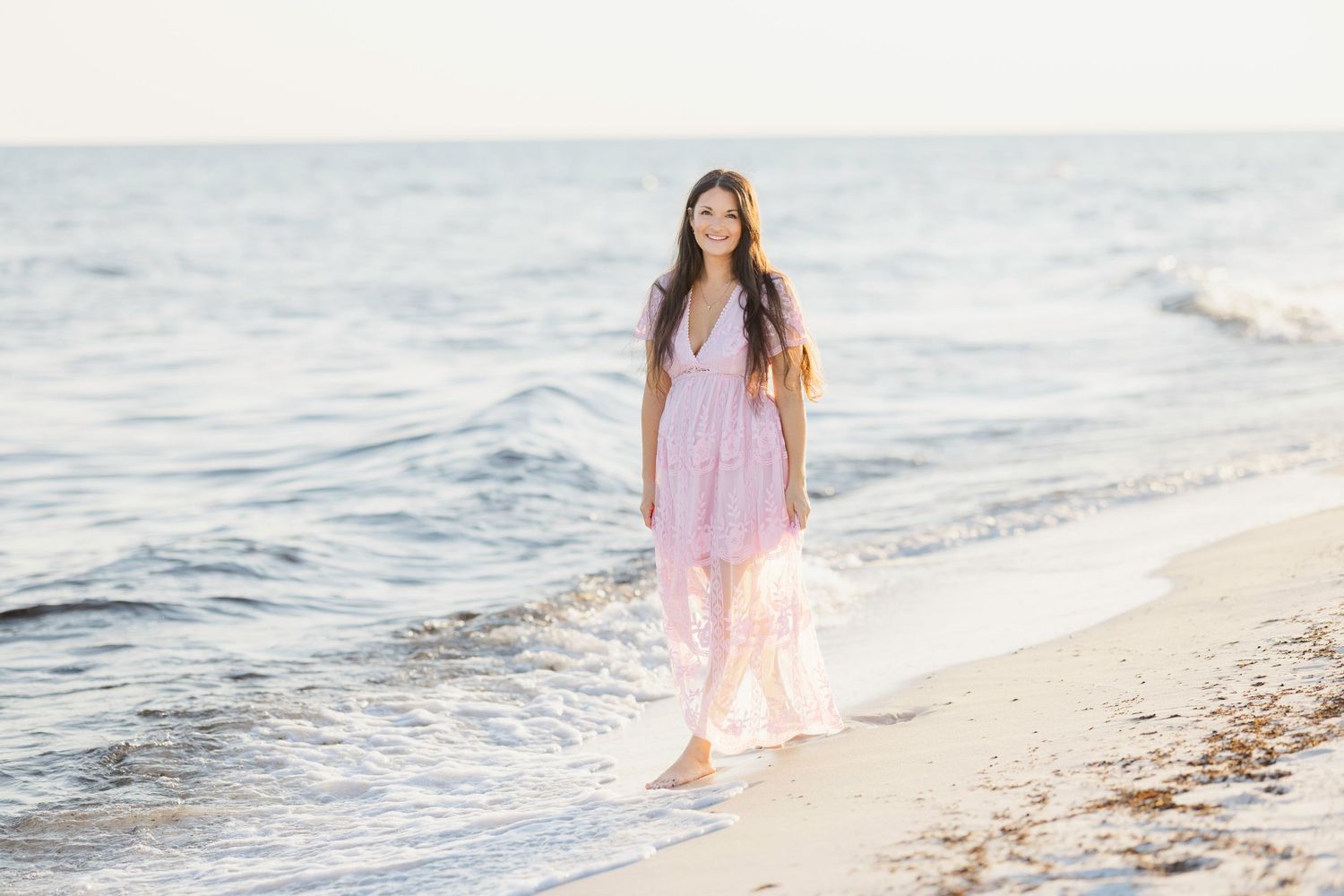 Young woman in pink maxi dress walking barefoot on sandy beach with ocean waves in background.