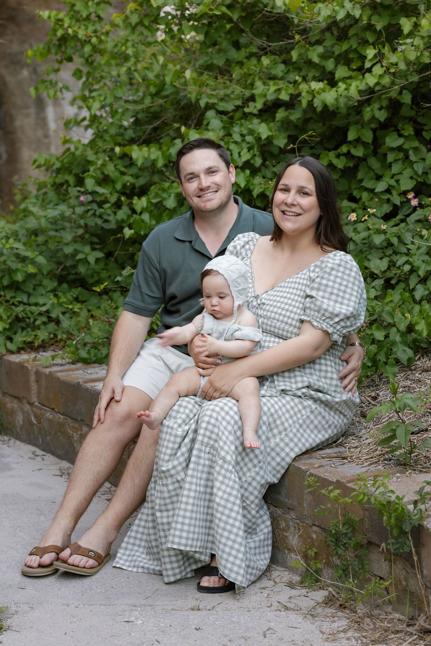 A smiling family of three sits outdoors on stone steps, parents in green outfits holding their baby in a bonnet.