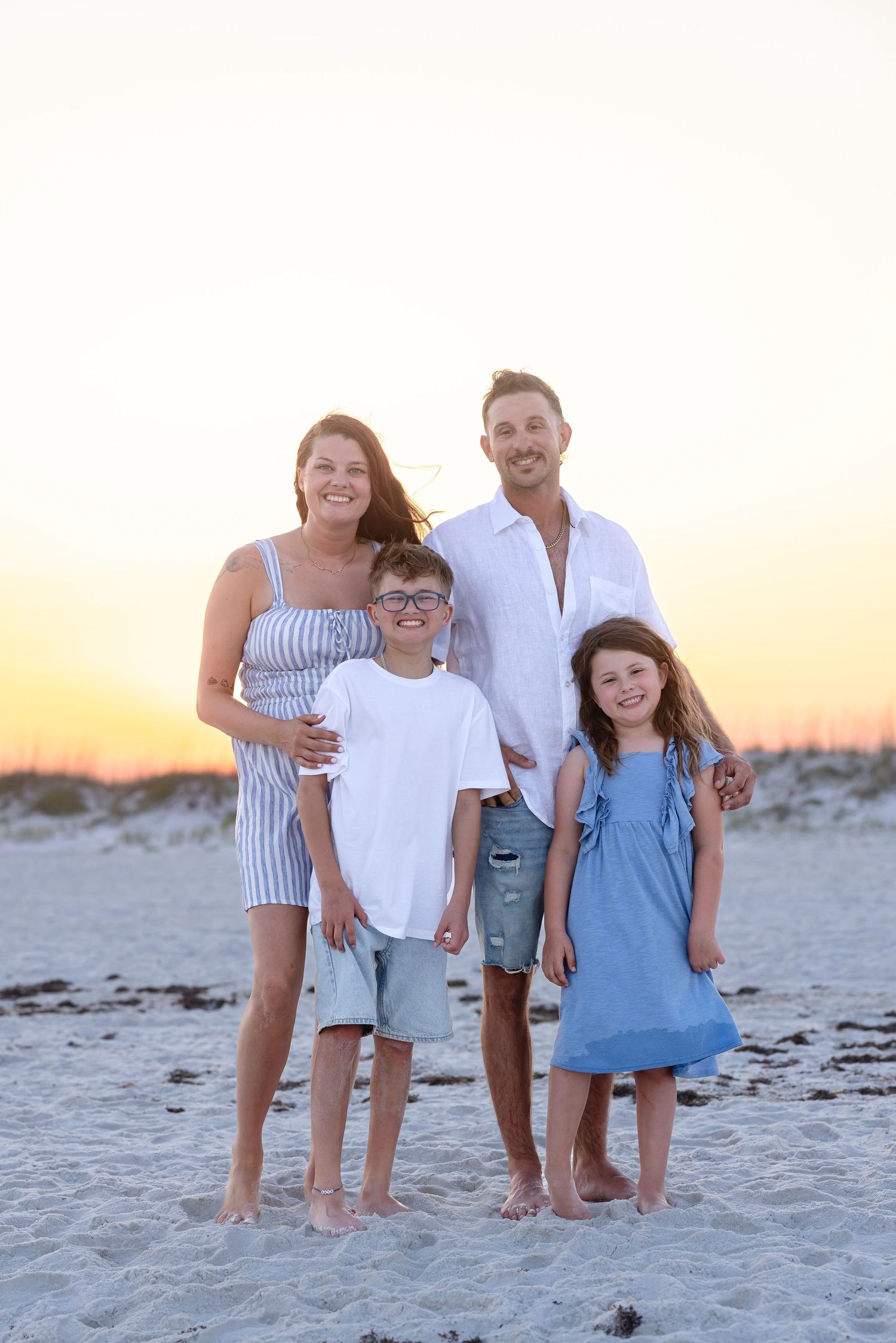 Happy family of four posing barefoot on beach at sunset, parents with two children in blue and white outfits.