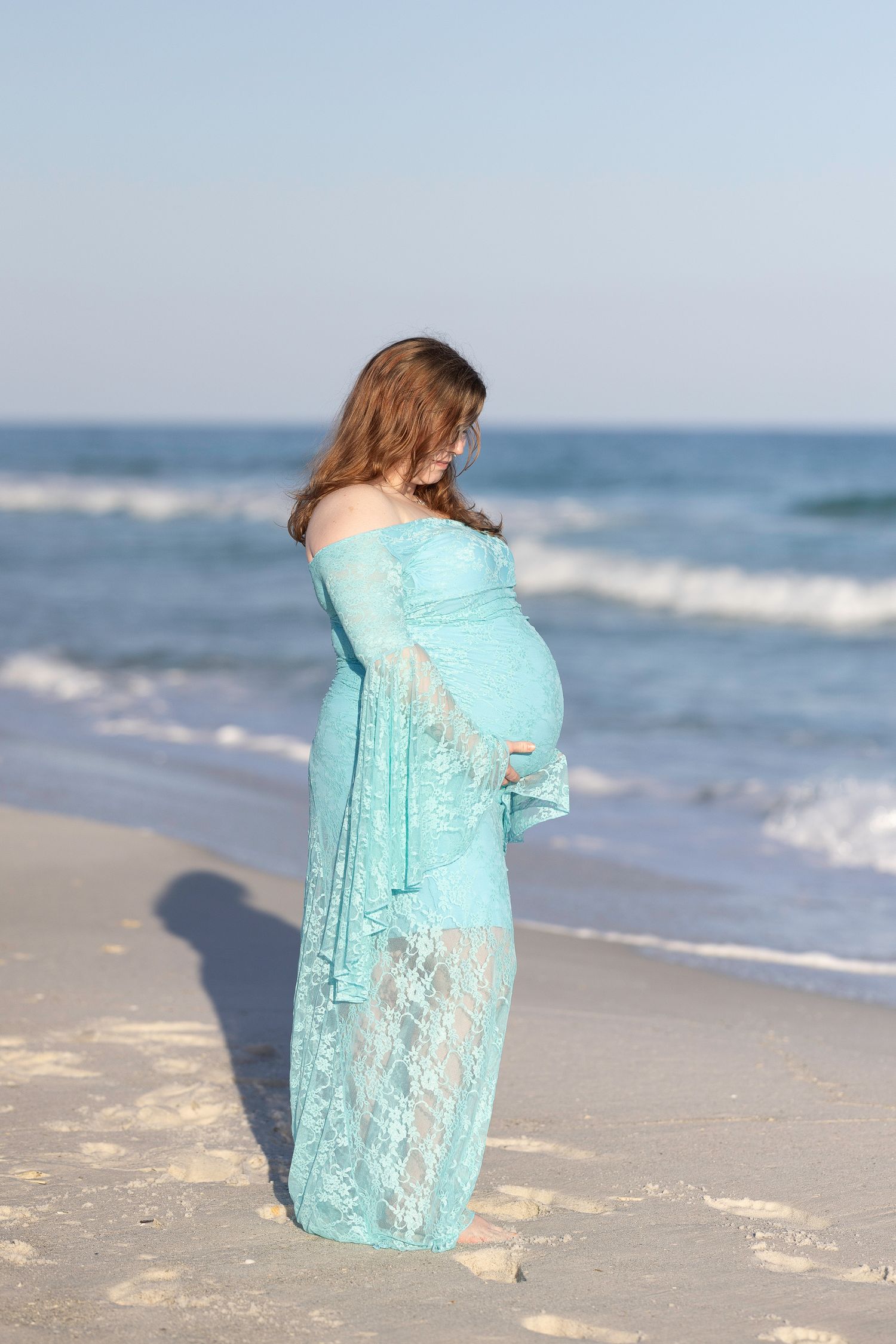 Pregnant woman in teal lace off-shoulder gown standing on sandy beach with ocean waves in background.