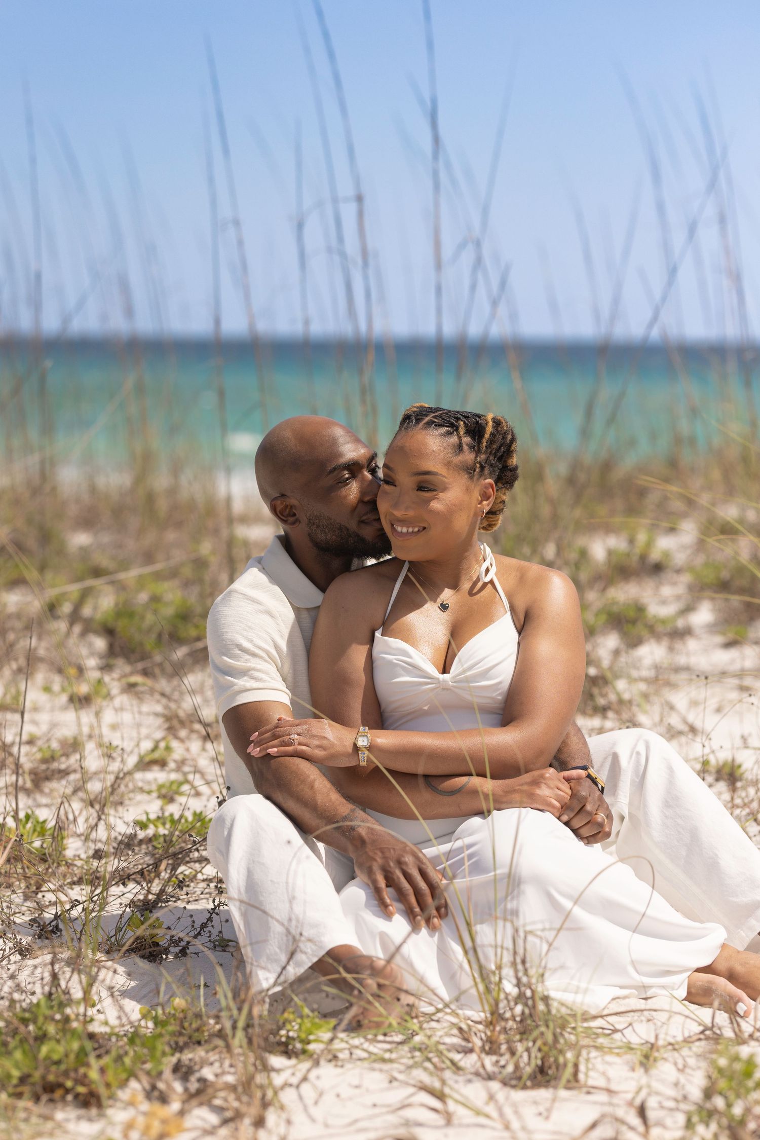 Romantic couple in white outfits sitting together on sandy beach dunes with turquoise ocean in background.