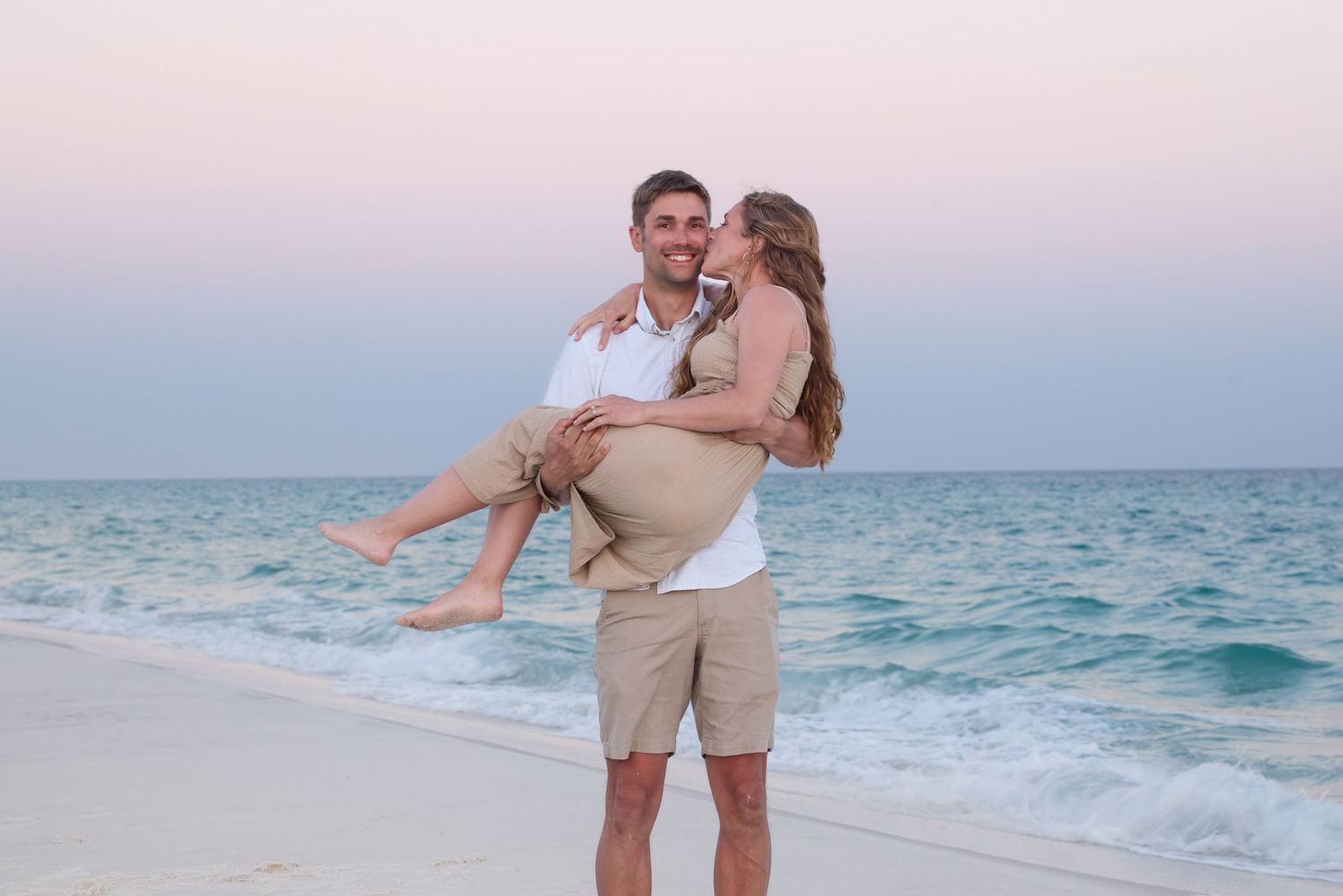 A man carrying a woman in his arms on a sandy beach at sunset, both smiling joyfully.