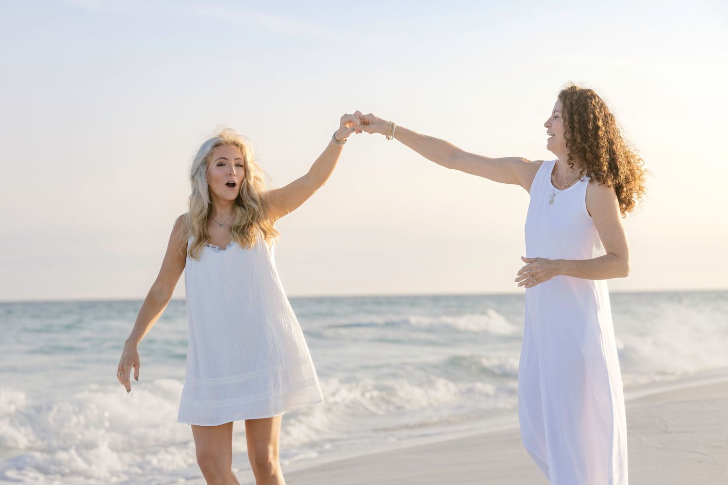 Two women in white dresses dancing joyfully on a sandy beach with ocean waves in the background.