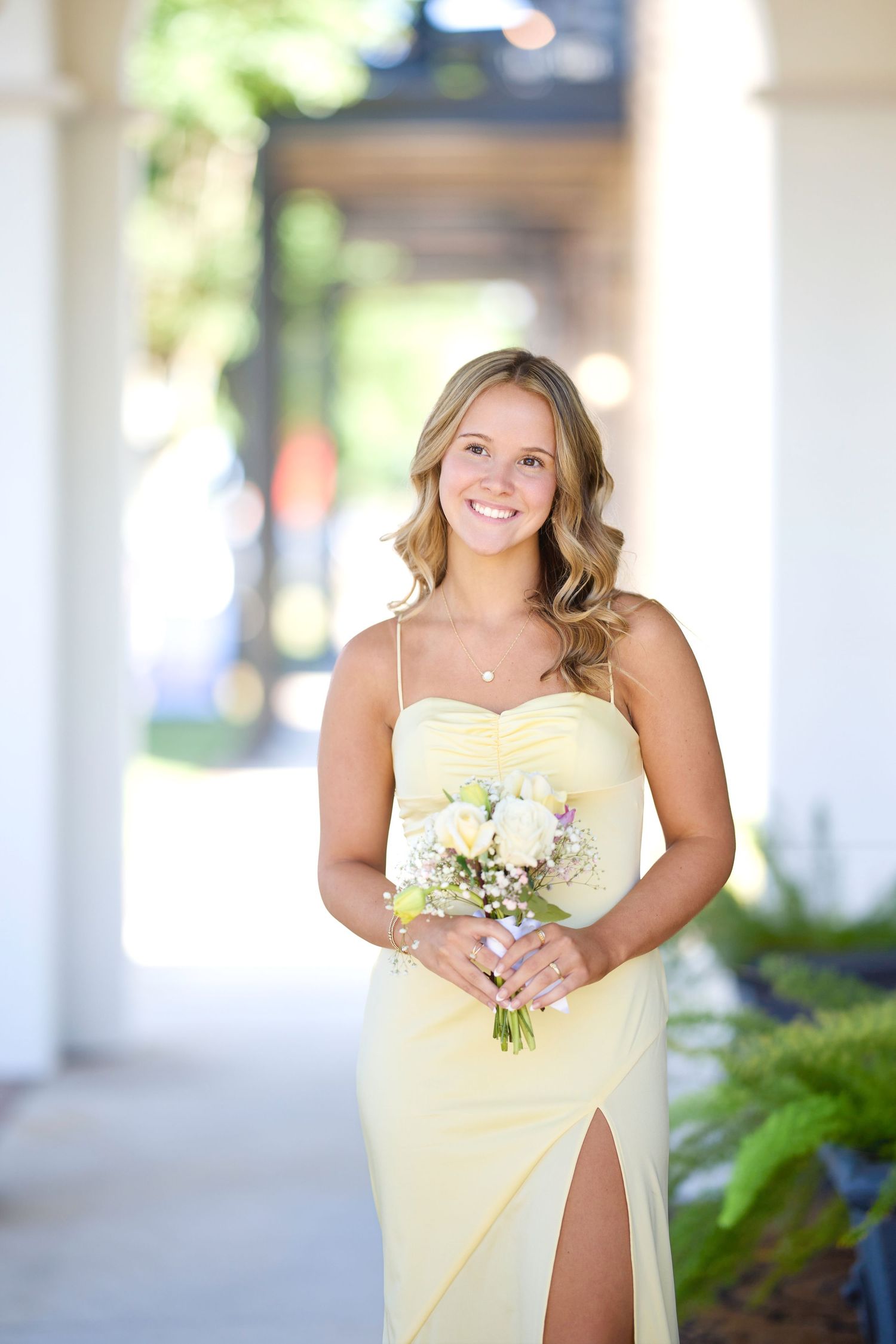 A bridesmaid in a pale yellow dress with a thigh slit holds a white floral bouquet while standing in a columned walkway.