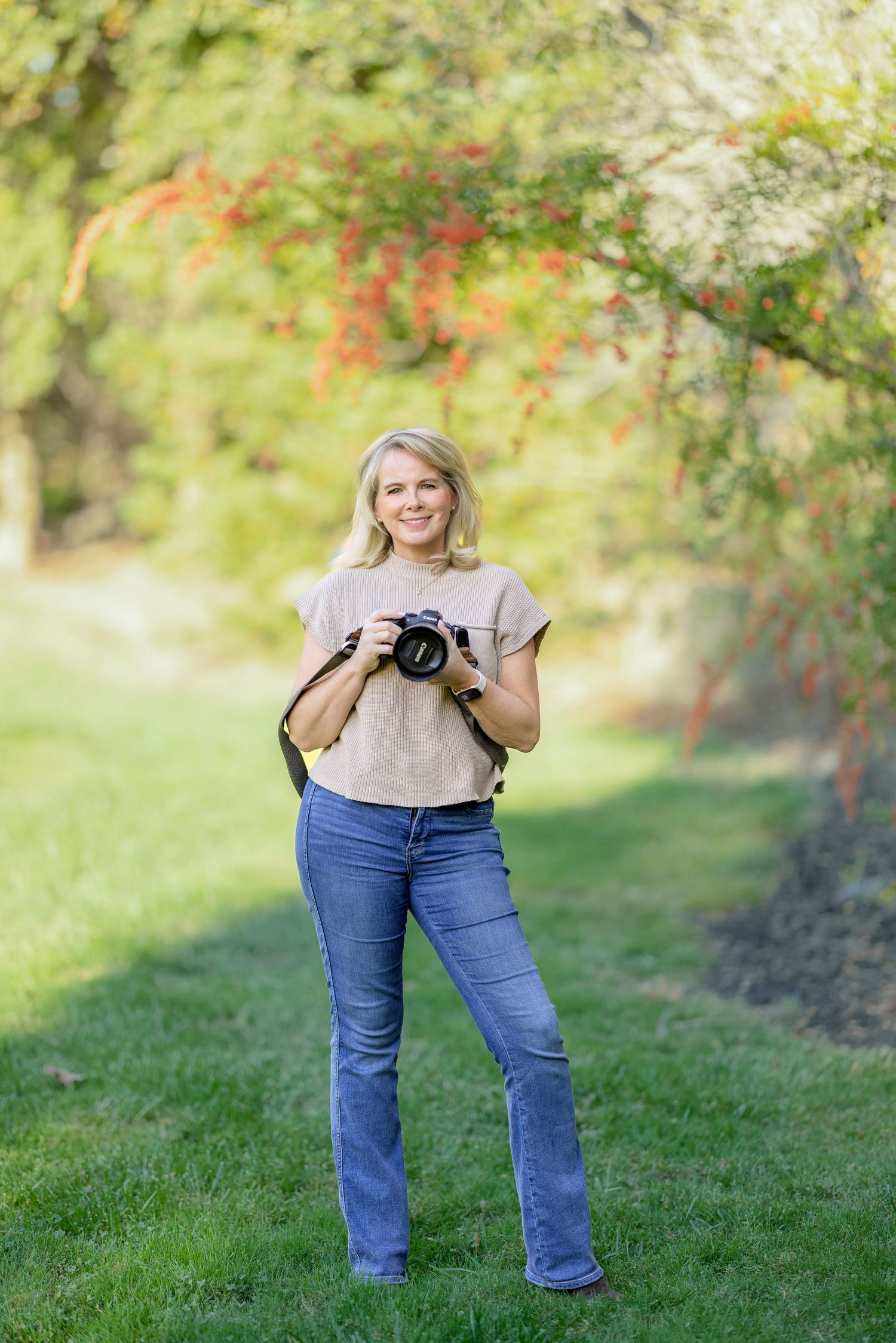 A photographer in jeans and beige sweater poses playfully in a grassy outdoor setting with fall foliage.
