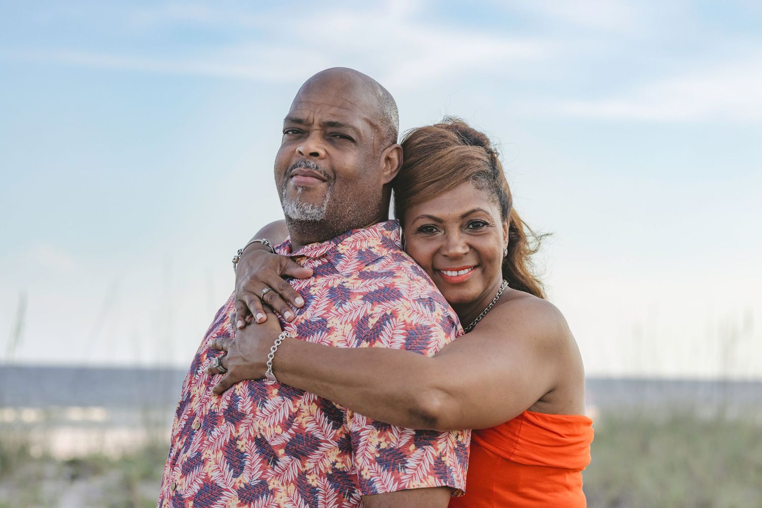 A happy couple embraces and smiles on a sunny beach, the man in a floral shirt and woman in an orange top.