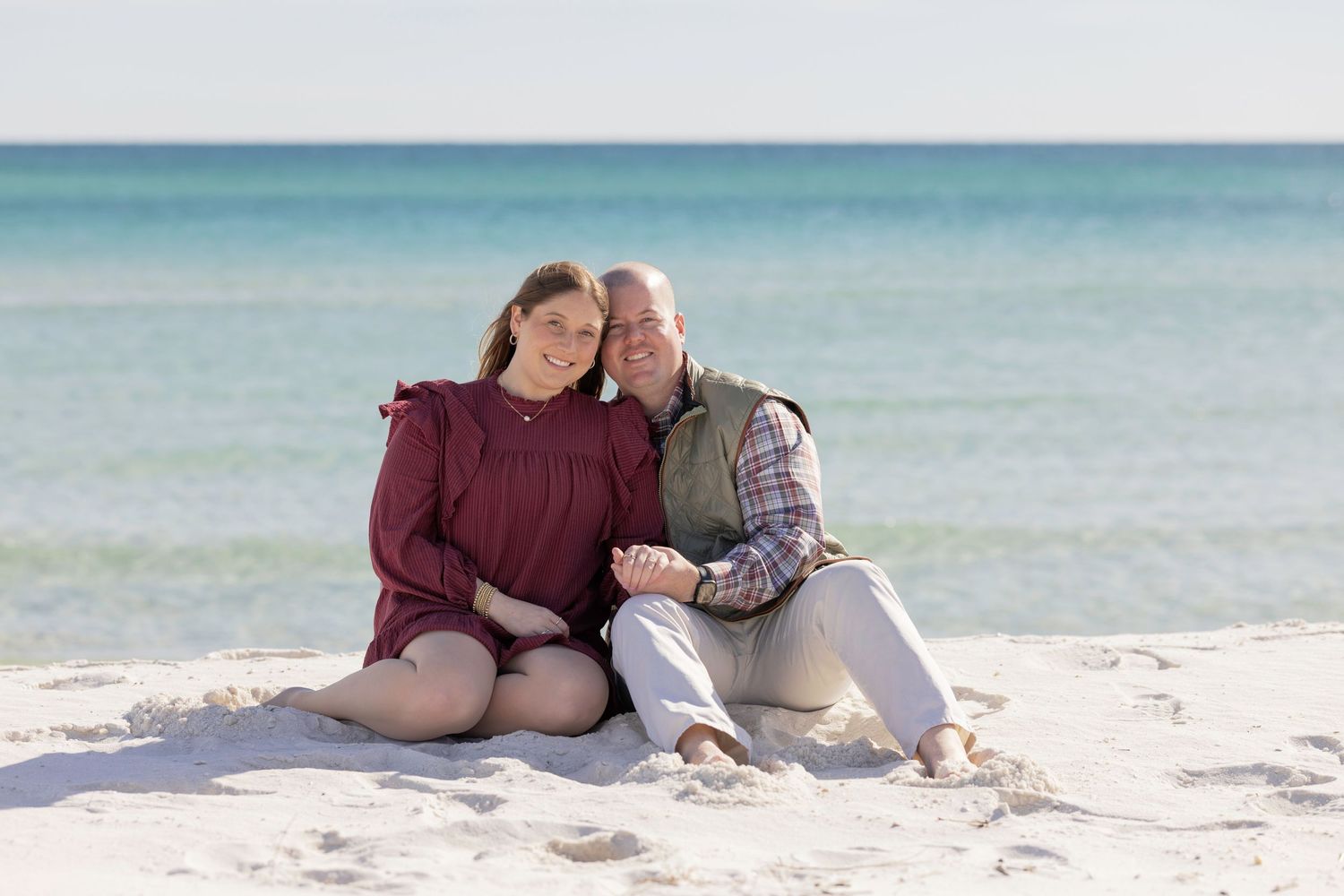 Couple sitting together on white sand beach, smiling, with turquoise ocean water in the background on a sunny day.