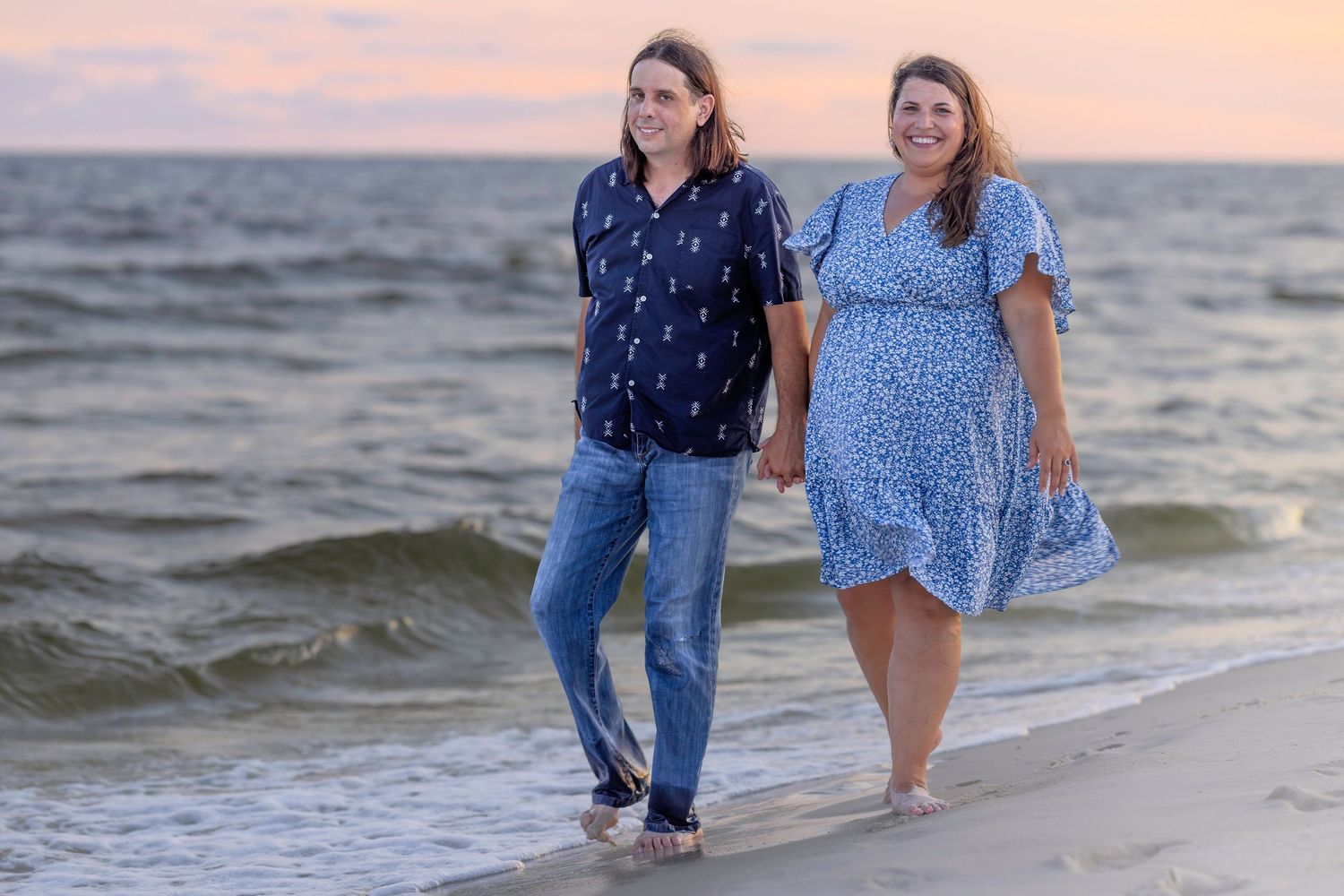 Happy couple walking hand in hand on beach at sunset, dressed in coordinating blue outfits.