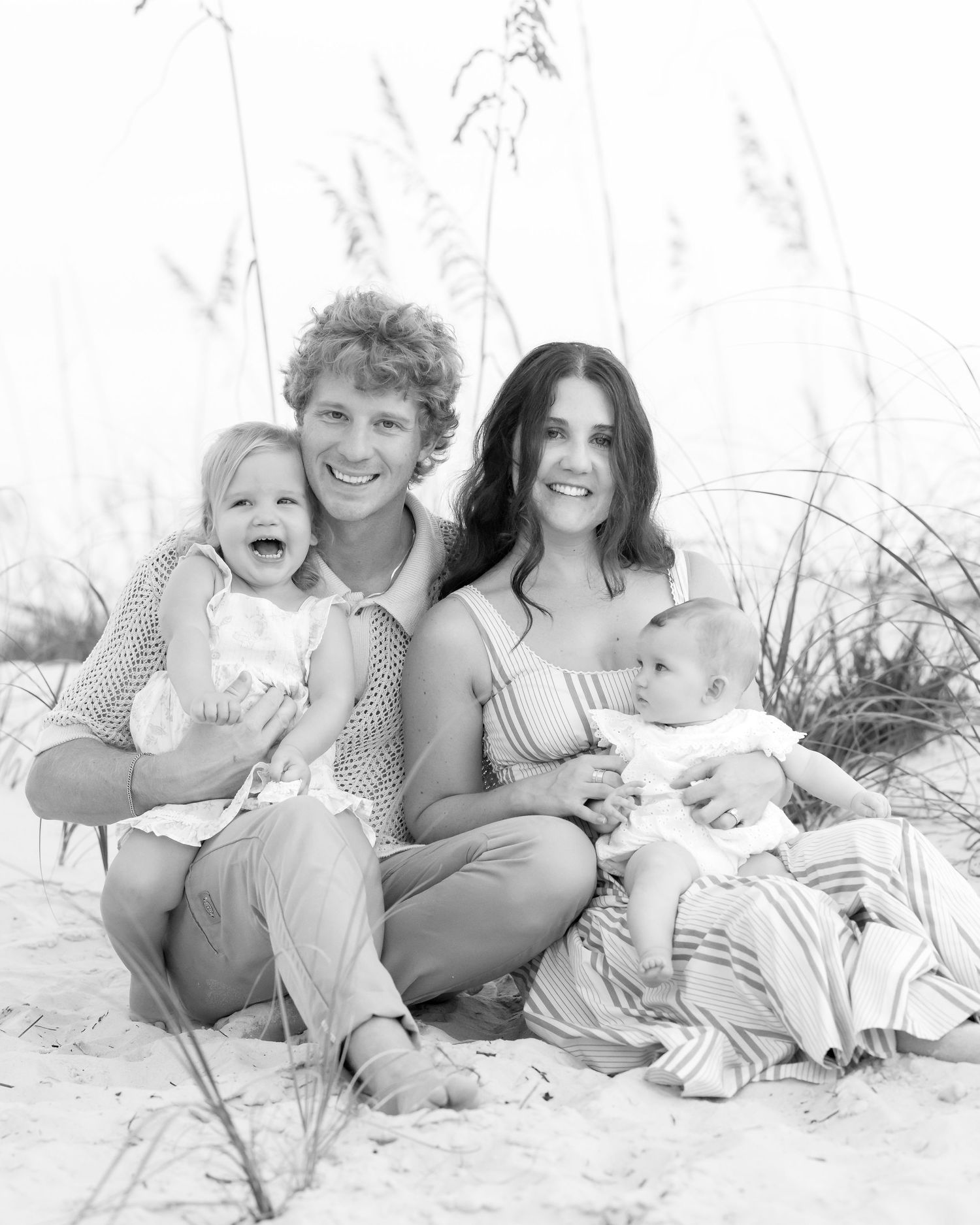 Black and white family portrait on beach with parents and two young children sitting in sand near sea oats.