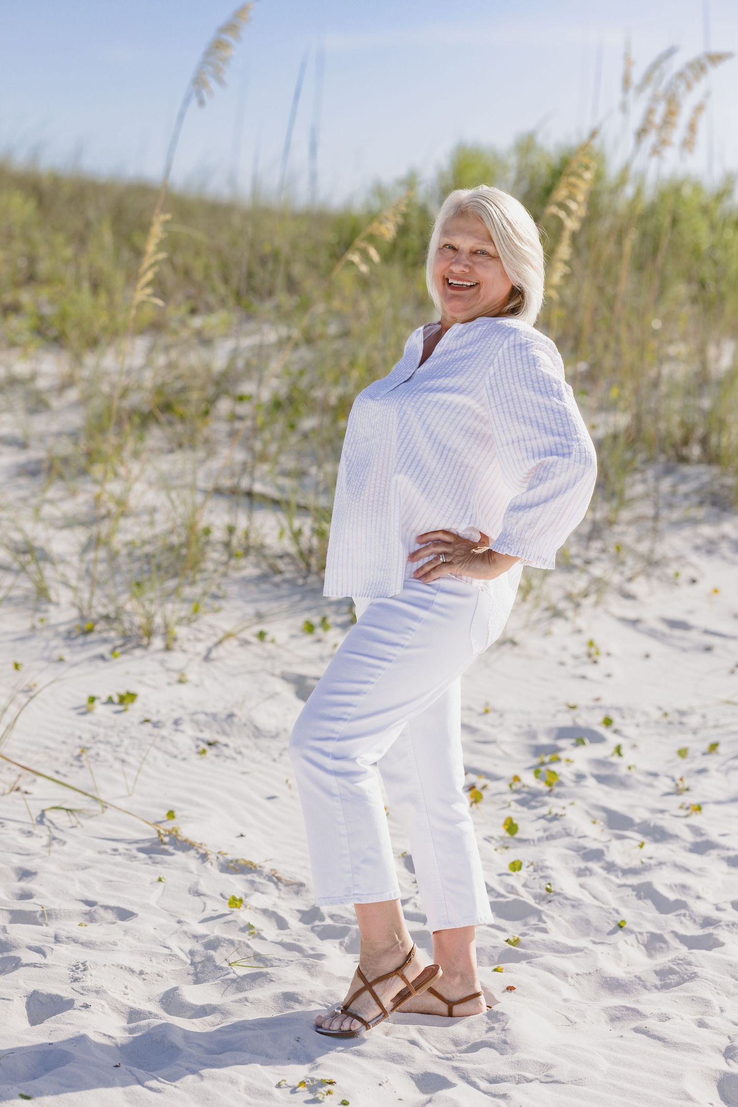 Smiling senior woman in white outfit and sandals posing on sandy beach dunes with sea oats in background.