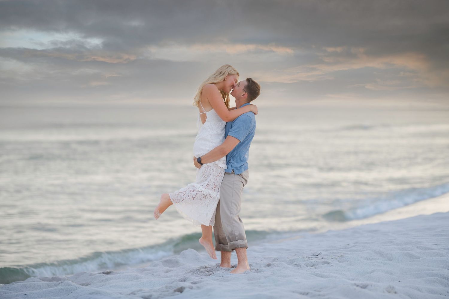 Couple sharing a romantic kiss on a sandy beach at sunset, with ocean waves and dramatic cloudy sky in background.