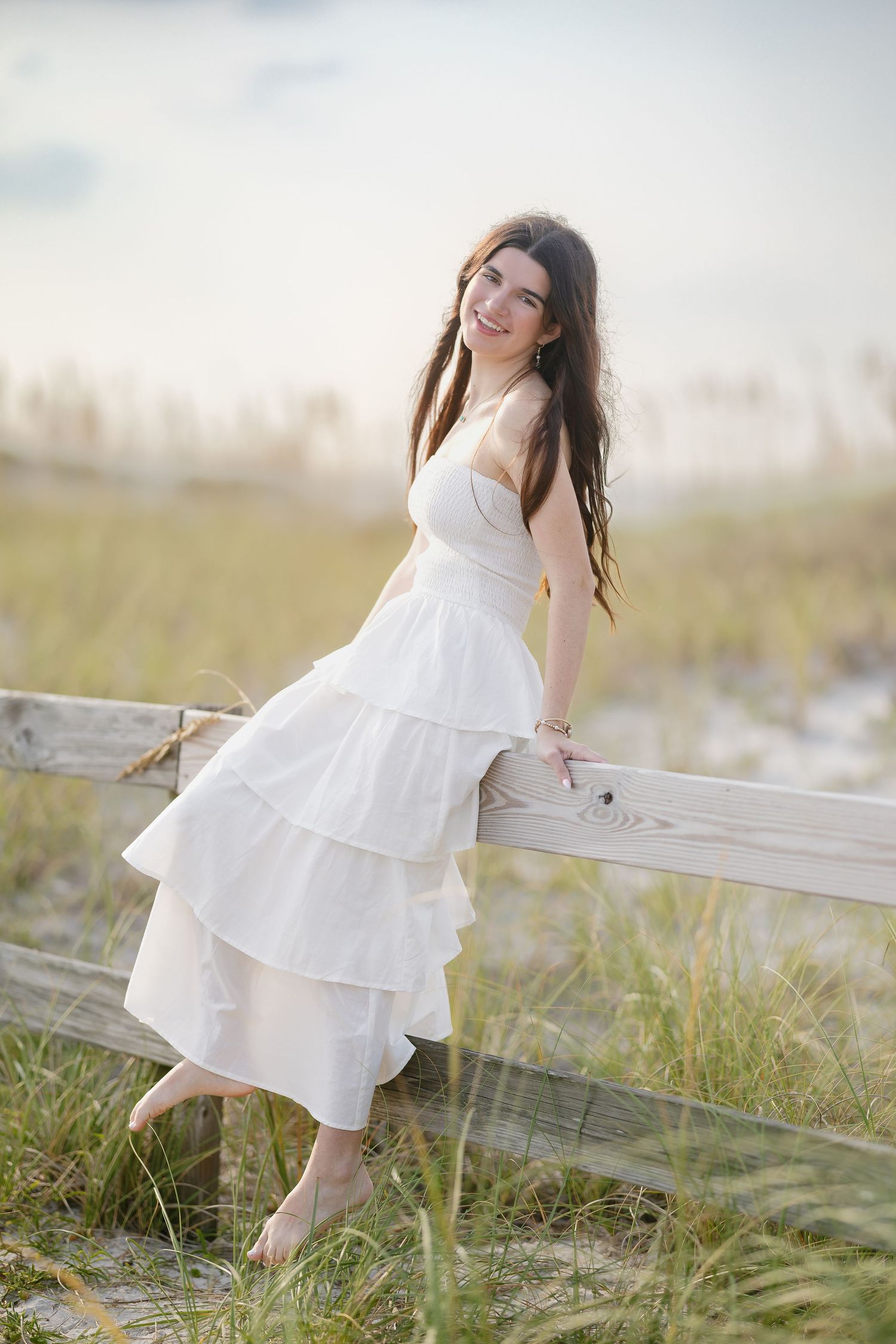 Young woman in white tiered dress leaning on wooden beach fence, smiling, with sandy dunes and grass background.