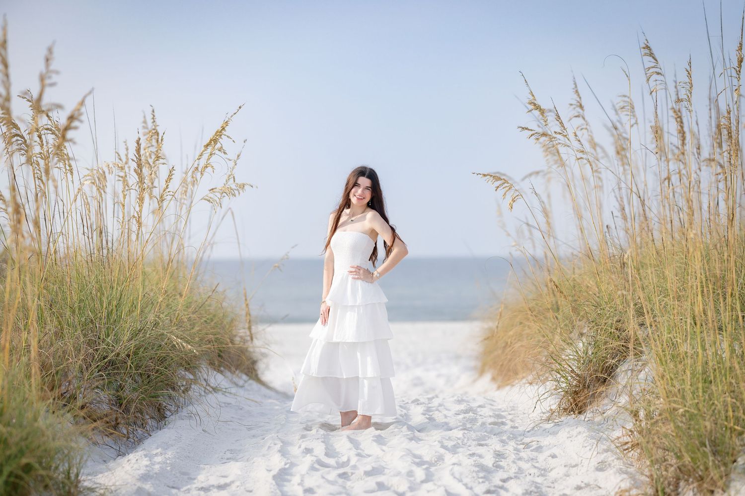 Brunette woman in white ruffled maxi dress posing on white sand beach path surrounded by tall sea oats.