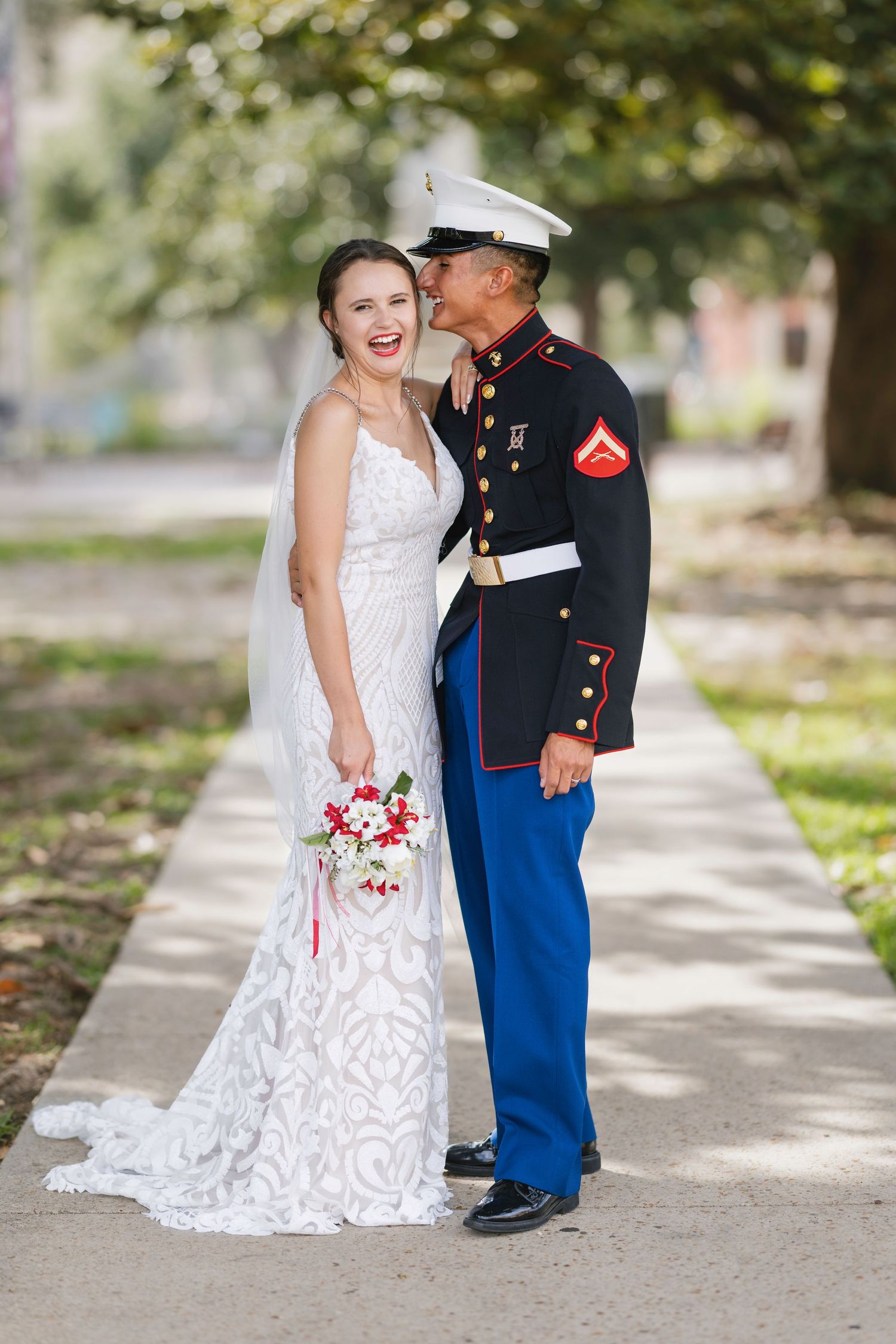 Marine groom kisses bride's cheek as she smiles holding red and white bouquet.