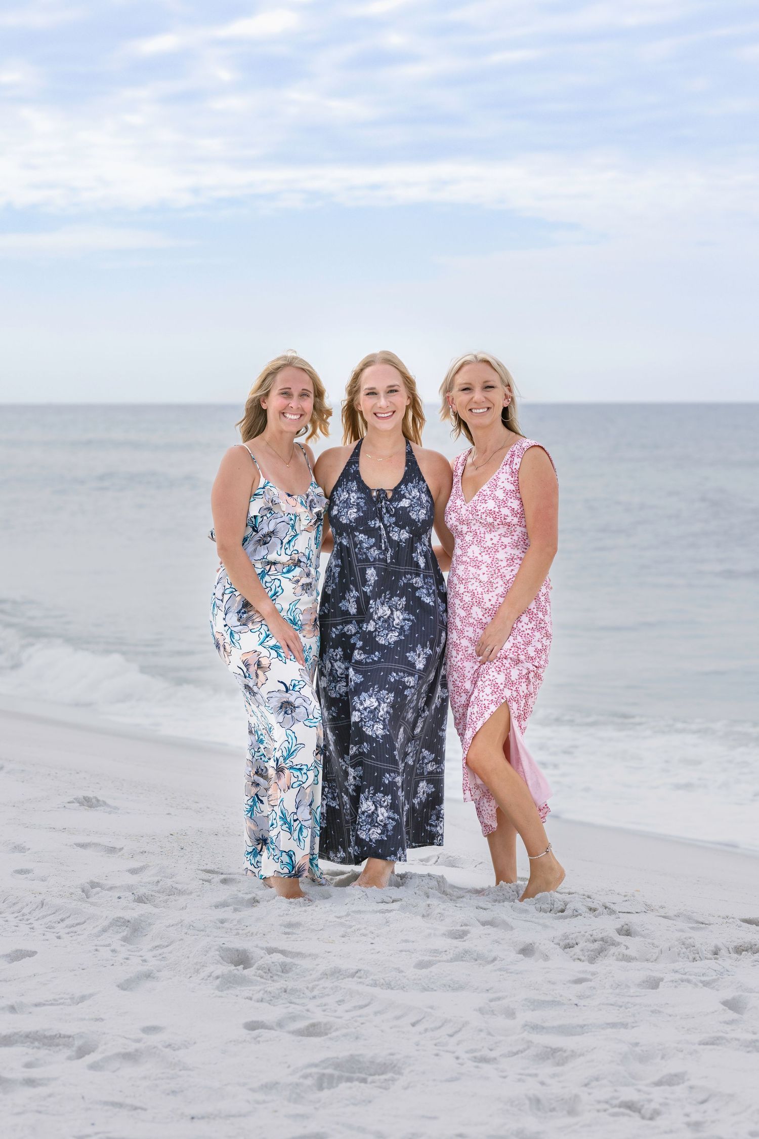 Three women in floral maxi dresses posing together on a sandy beach with ocean waves behind them.