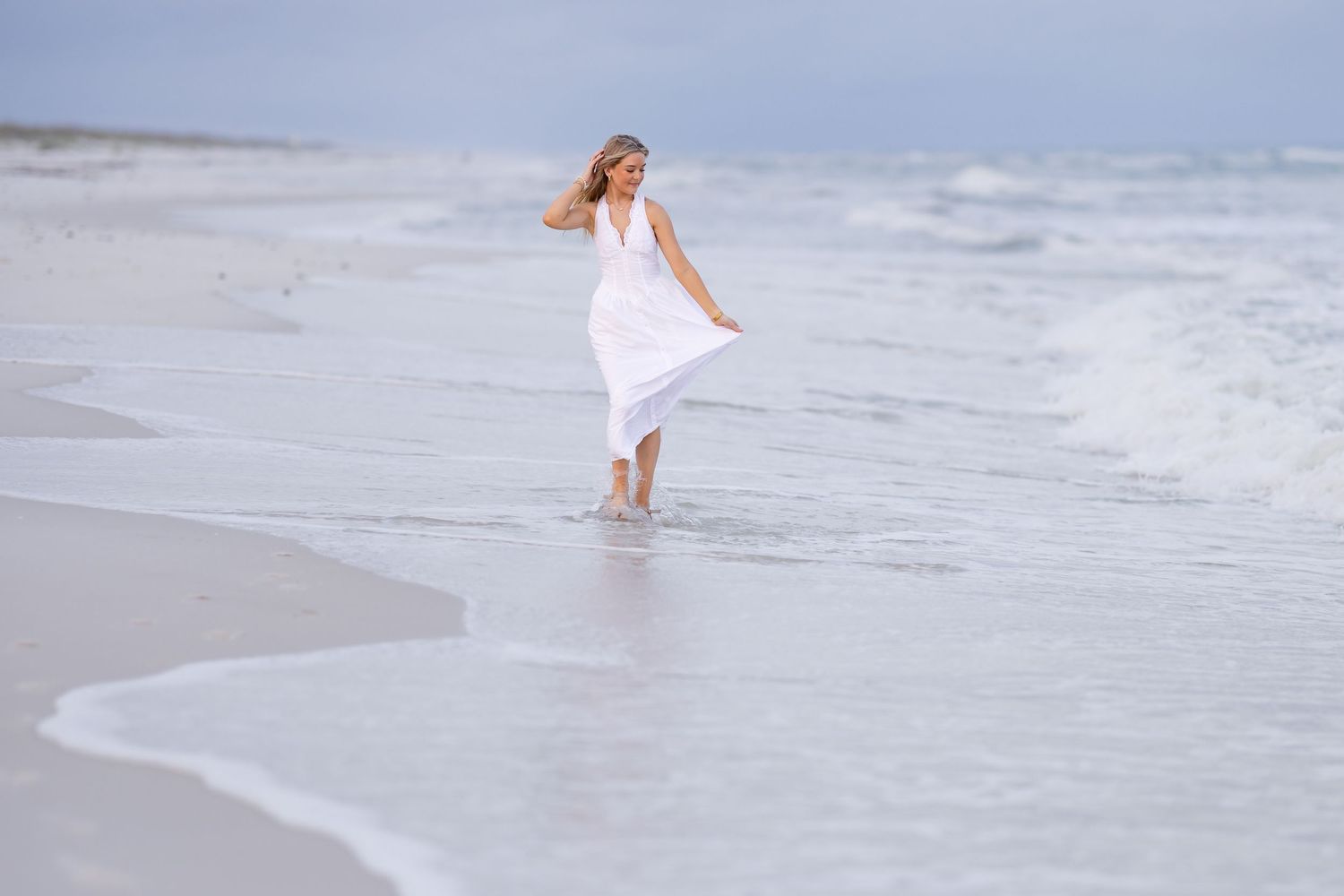 Woman in white dress walking along sandy beach shoreline with ocean waves, enjoying a serene coastal day.