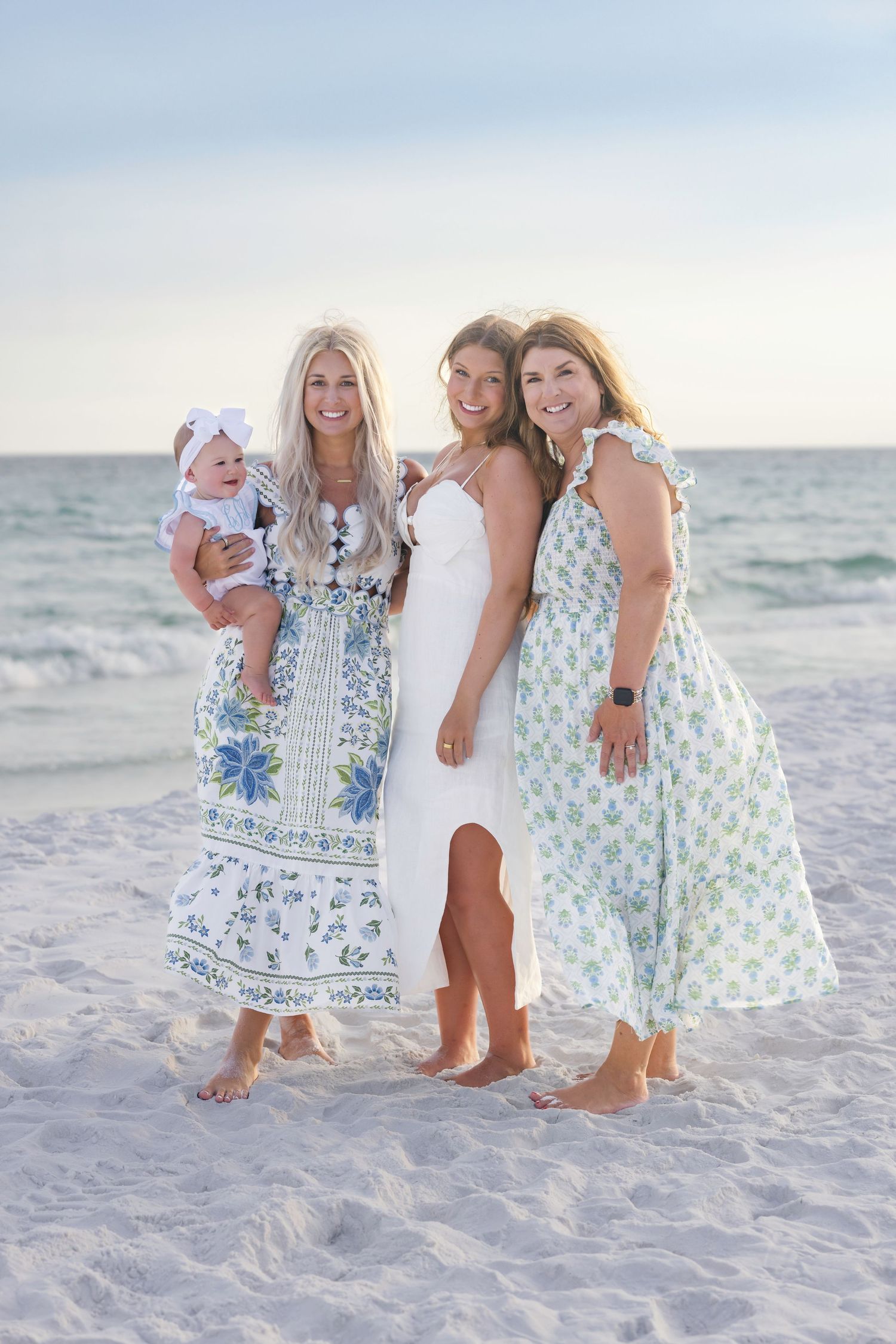 Three women and a baby posing on a sandy beach in floral and white dresses at sunset.