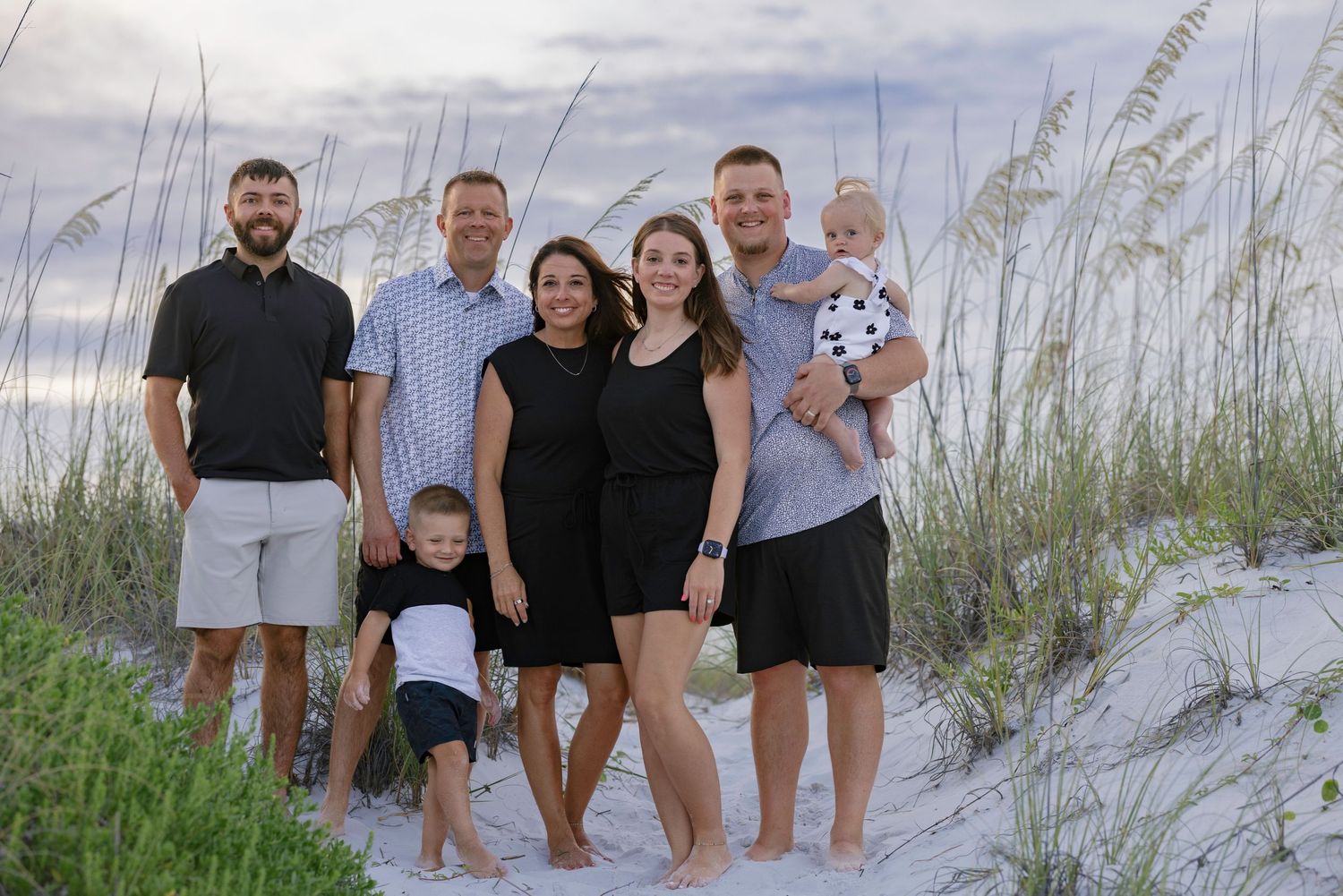 Extended family of seven poses together on white sand beach dunes at sunset near sea oats.