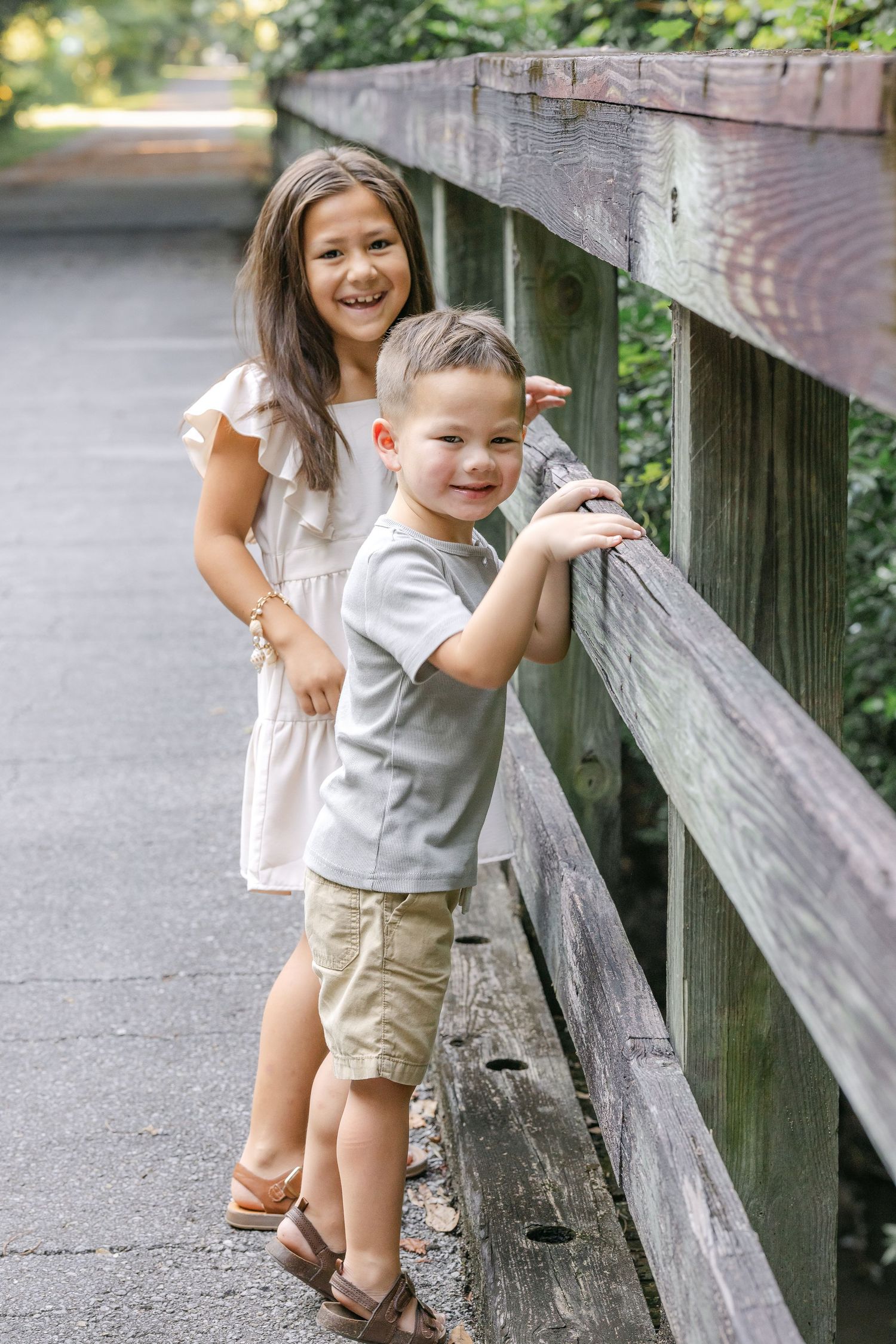Children pose playfully by a wooden railing on an outdoor path with greenery in the background.