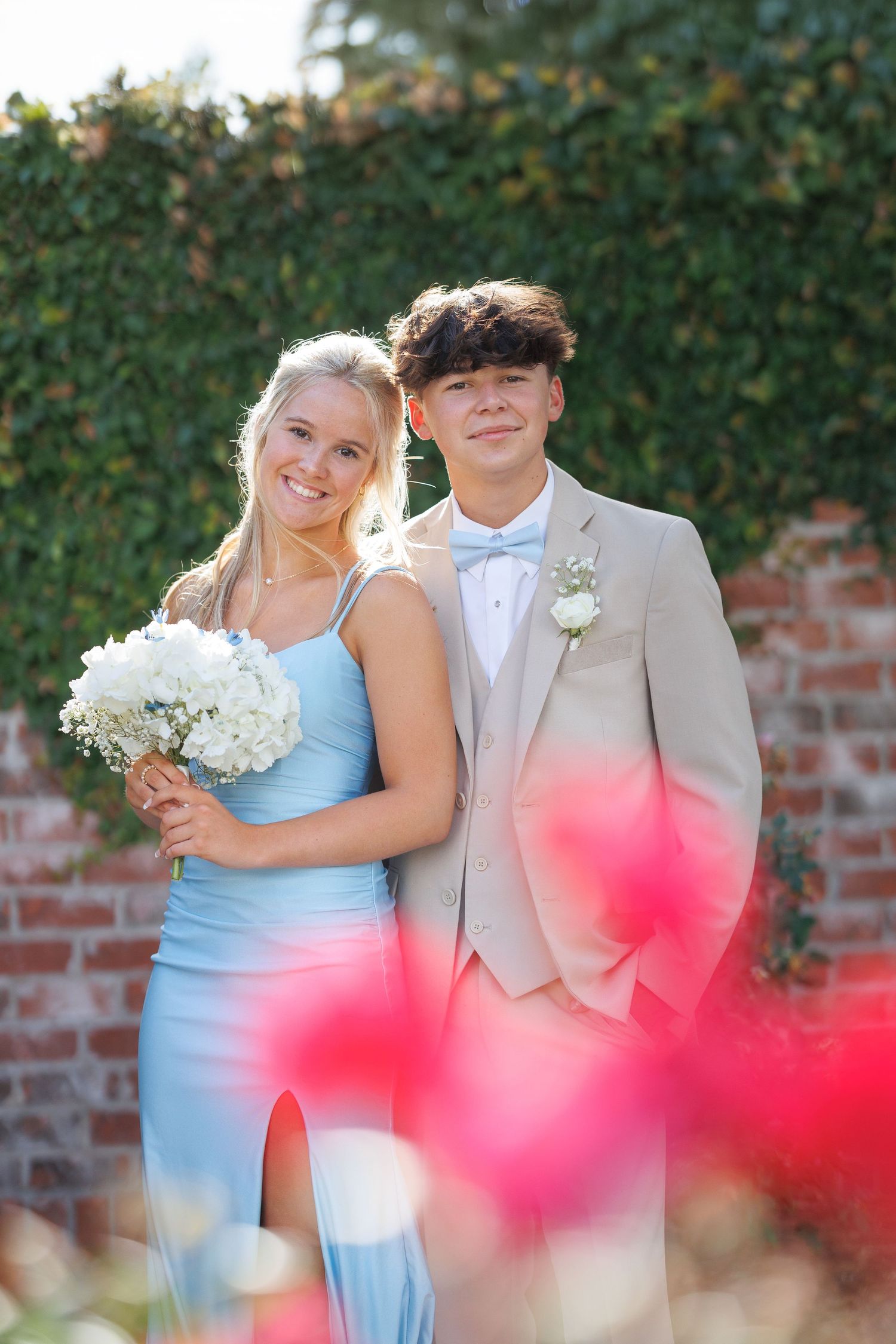 Young couple poses for formal portraits in front of brick wall with pink flowers in foreground.