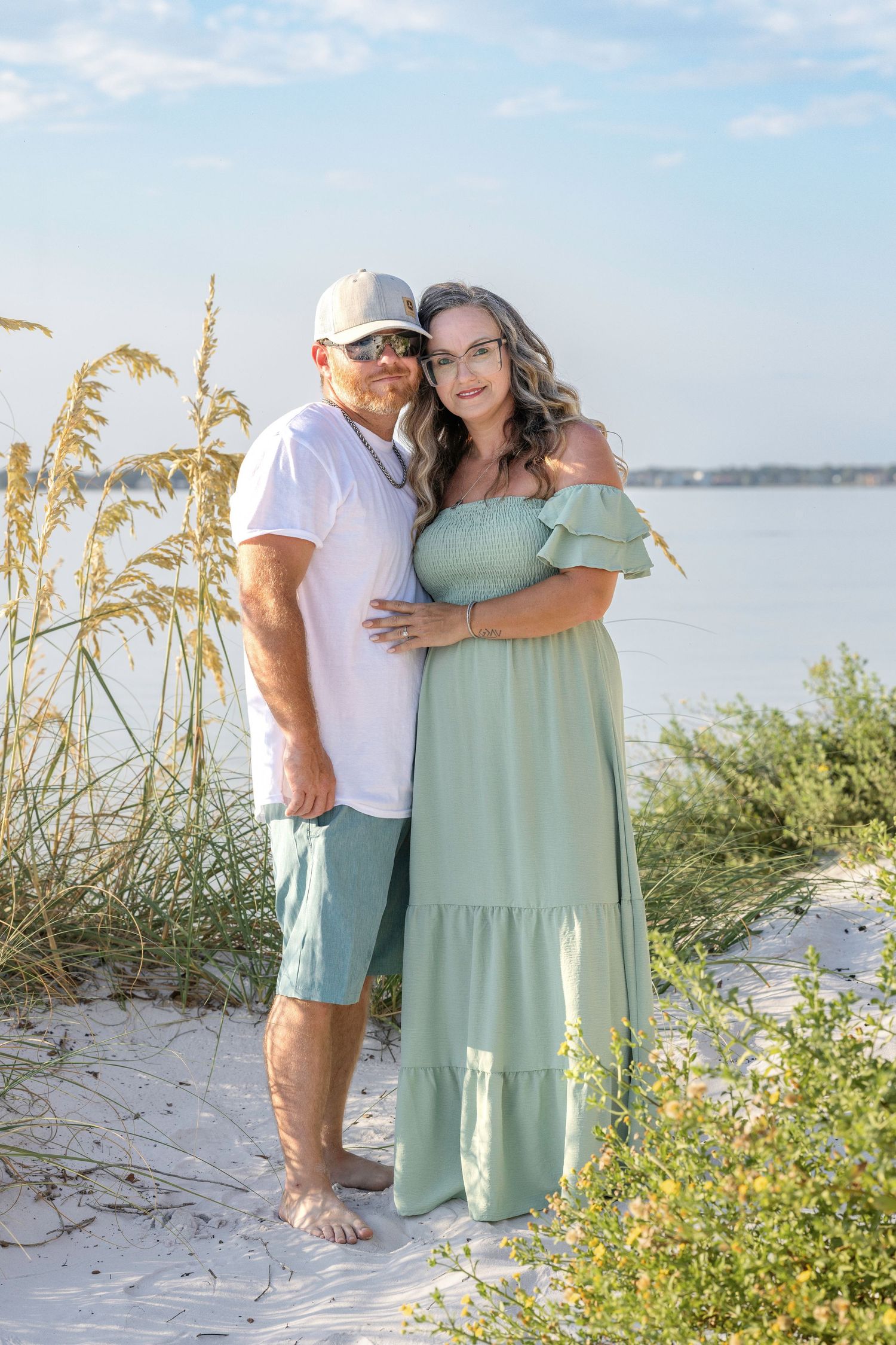 A couple poses together on a sandy beach with sea oats and water in the background on a sunny day.