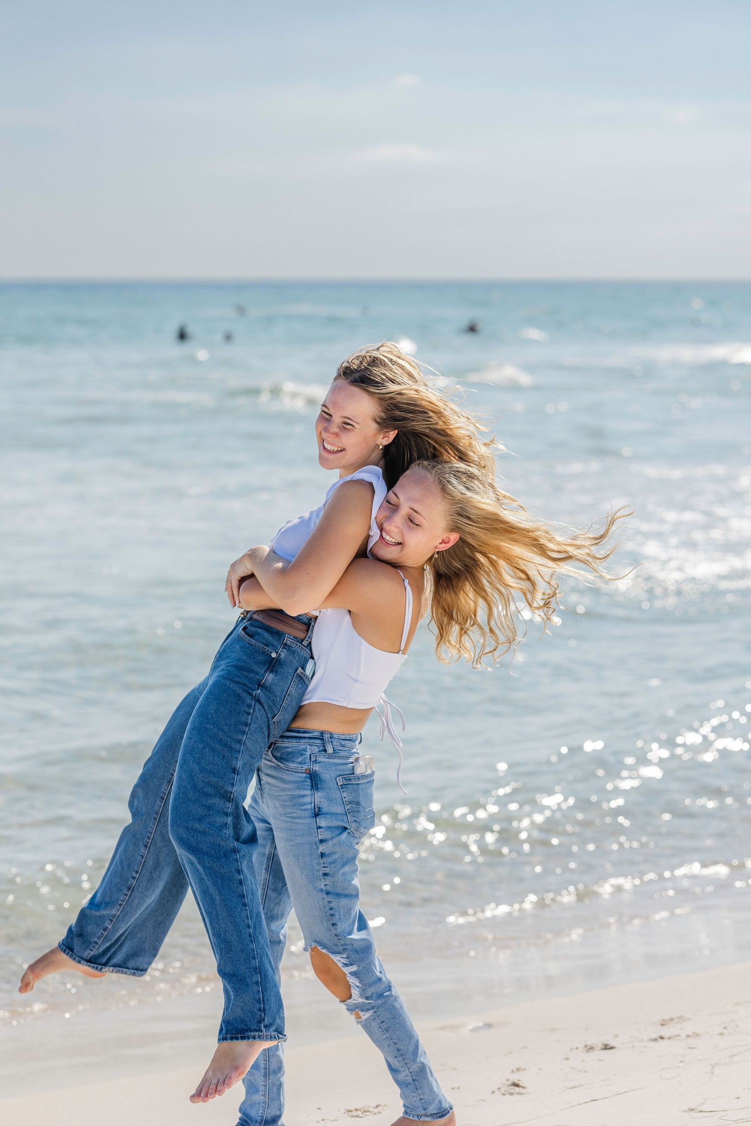Two friends in casual denim outfits dance and laugh together on a sunny beach with sparkling ocean waves in the background.