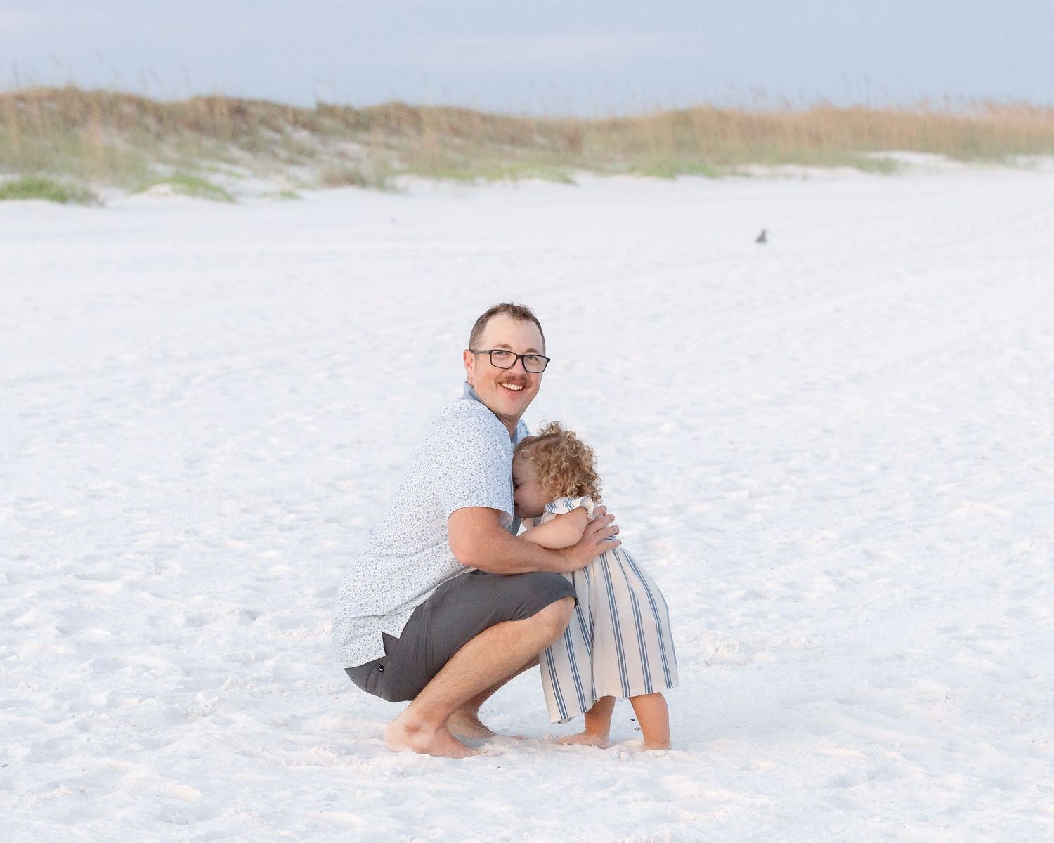 A father kneels on white sand beach hugging his young child, with sand dunes and sea grass in the background.