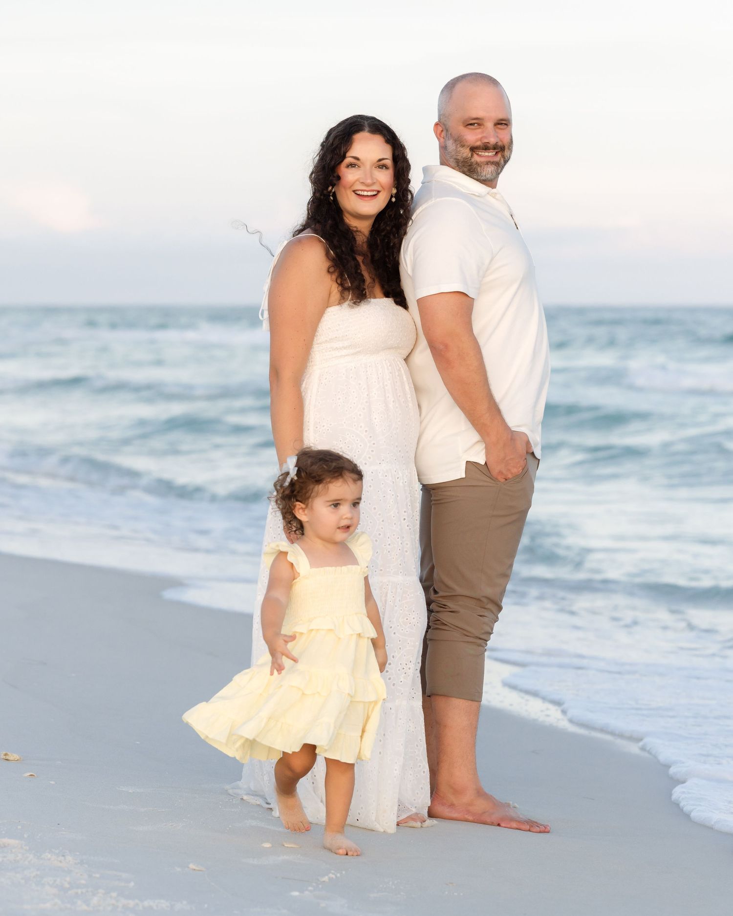 Family portrait on beach with couple in white standing back-to-back, toddler girl in yellow dress.