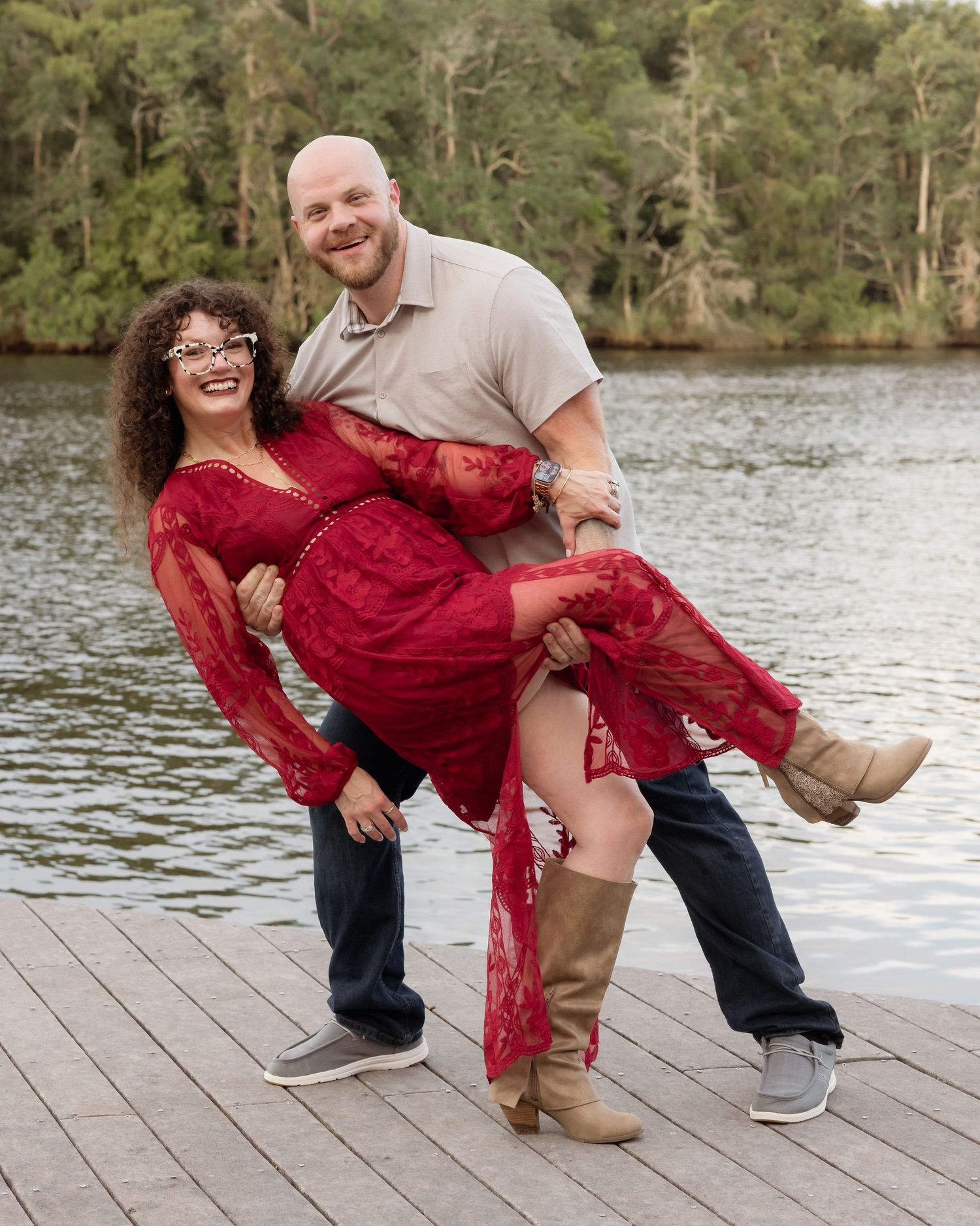 A smiling couple poses playfully on a wooden dock, man lifting woman in a red lace dress.