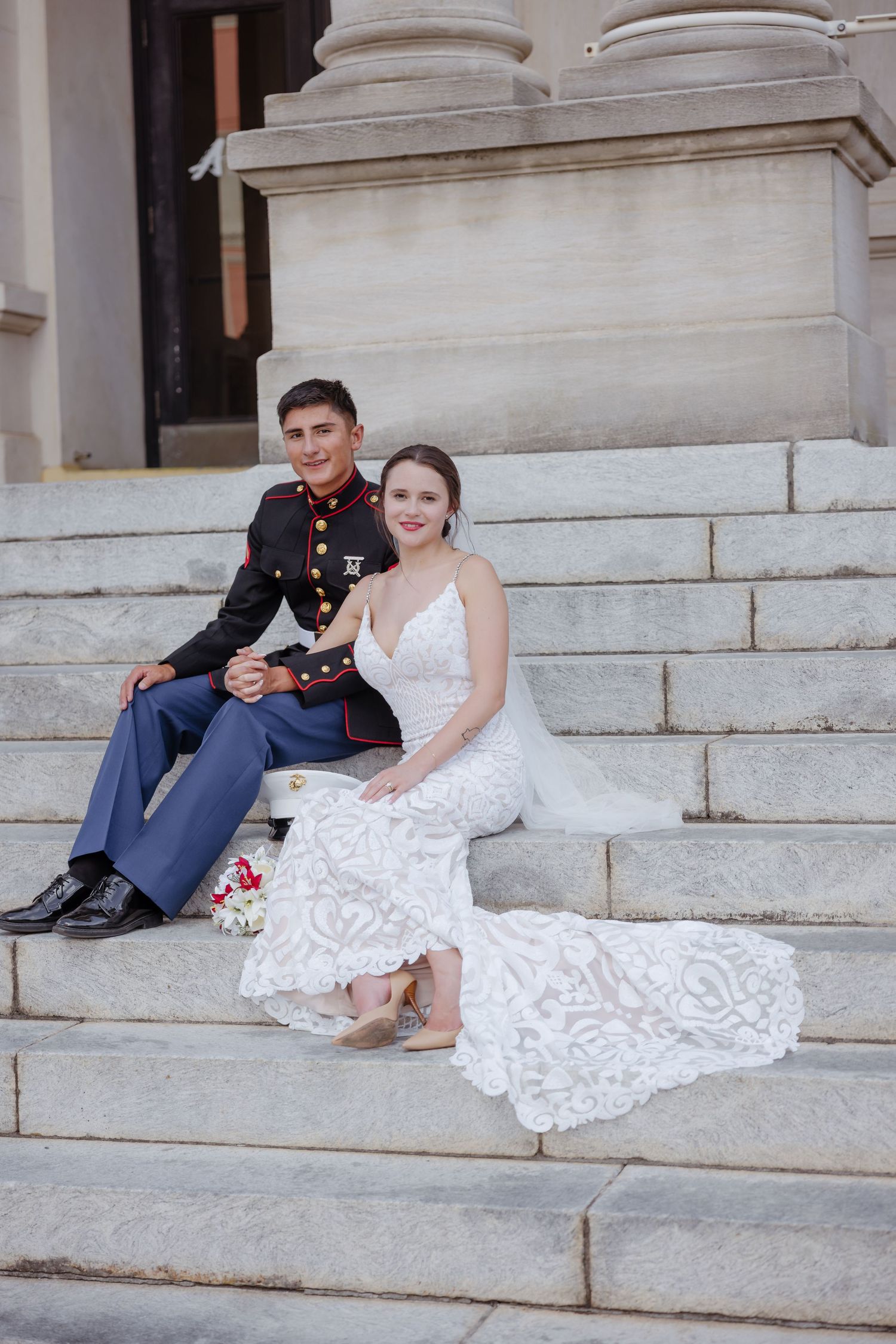 A Marine in dress uniform and bride in white lace gown sit together on marble steps for their wedding portrait.