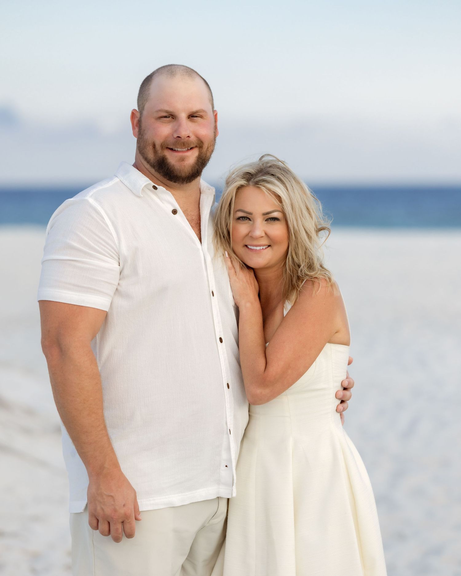 A smiling couple poses together on a white sand beach, both dressed in white outfits with the ocean in the background.