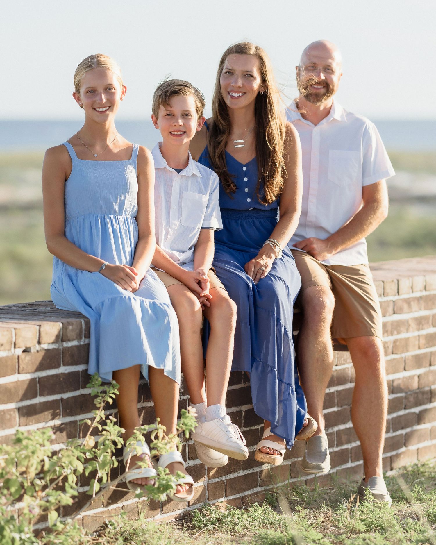 Family of four smiling and sitting on a brick wall outdoors near a beach on a sunny day.