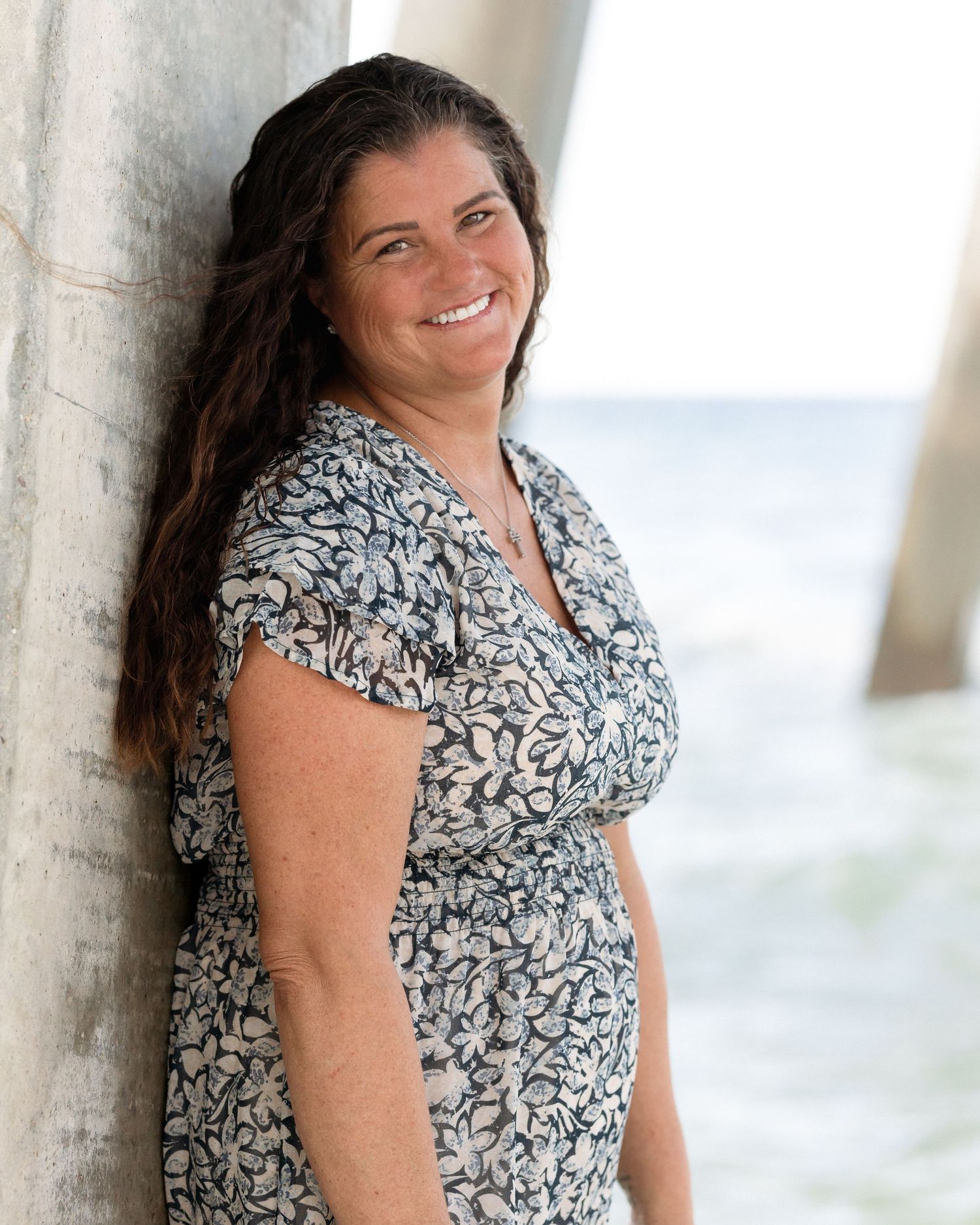 Smiling woman with long brown hair leaning against pier column in floral dress at the beach.