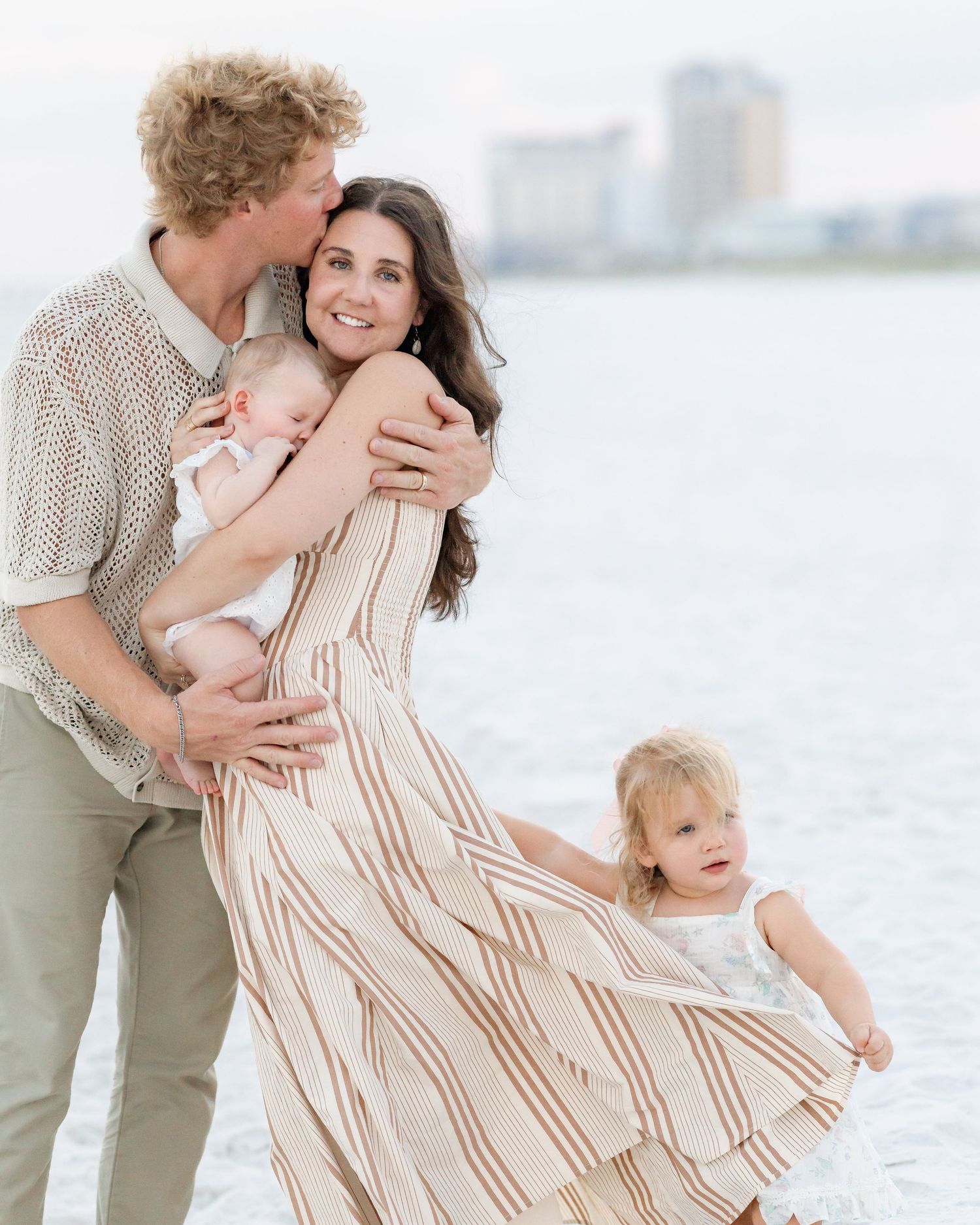 Loving family of four at beach, dad kissing mom while she holds newborn and toddler clings to her dress.