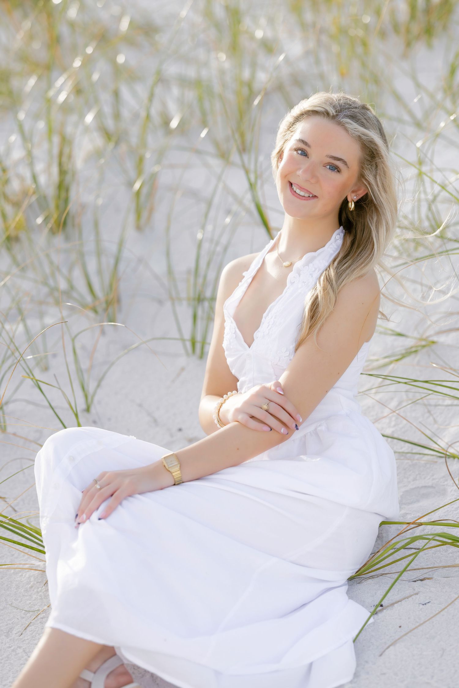 Graduate in white halter dress poses sitting on sand dunes with beach grass in soft sunlight.