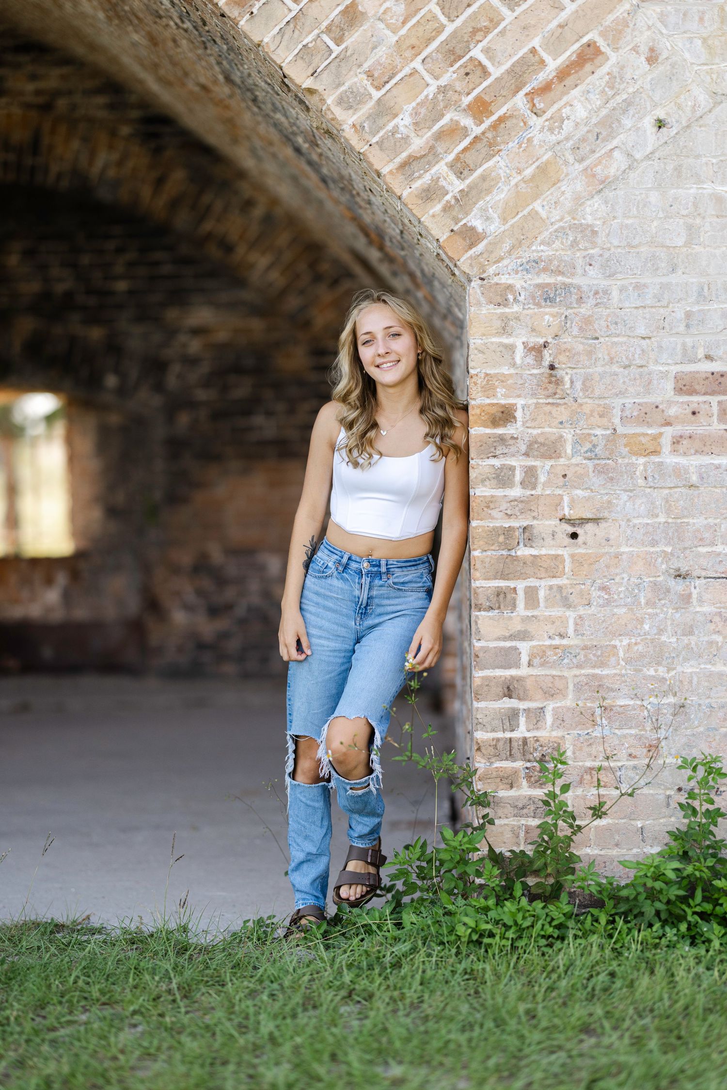 A person in a white top and blue jeans sits casually against a brick wall under an archway.