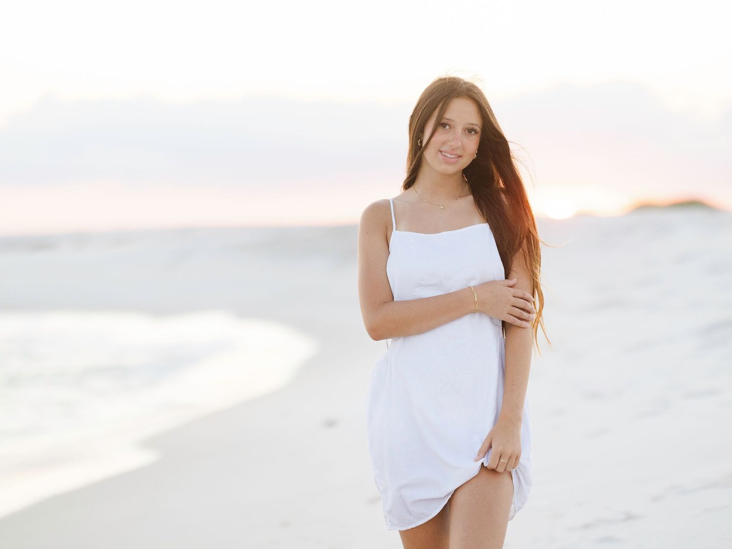 Woman in white swimsuit poses gracefully on sandy beach during golden sunset hour.