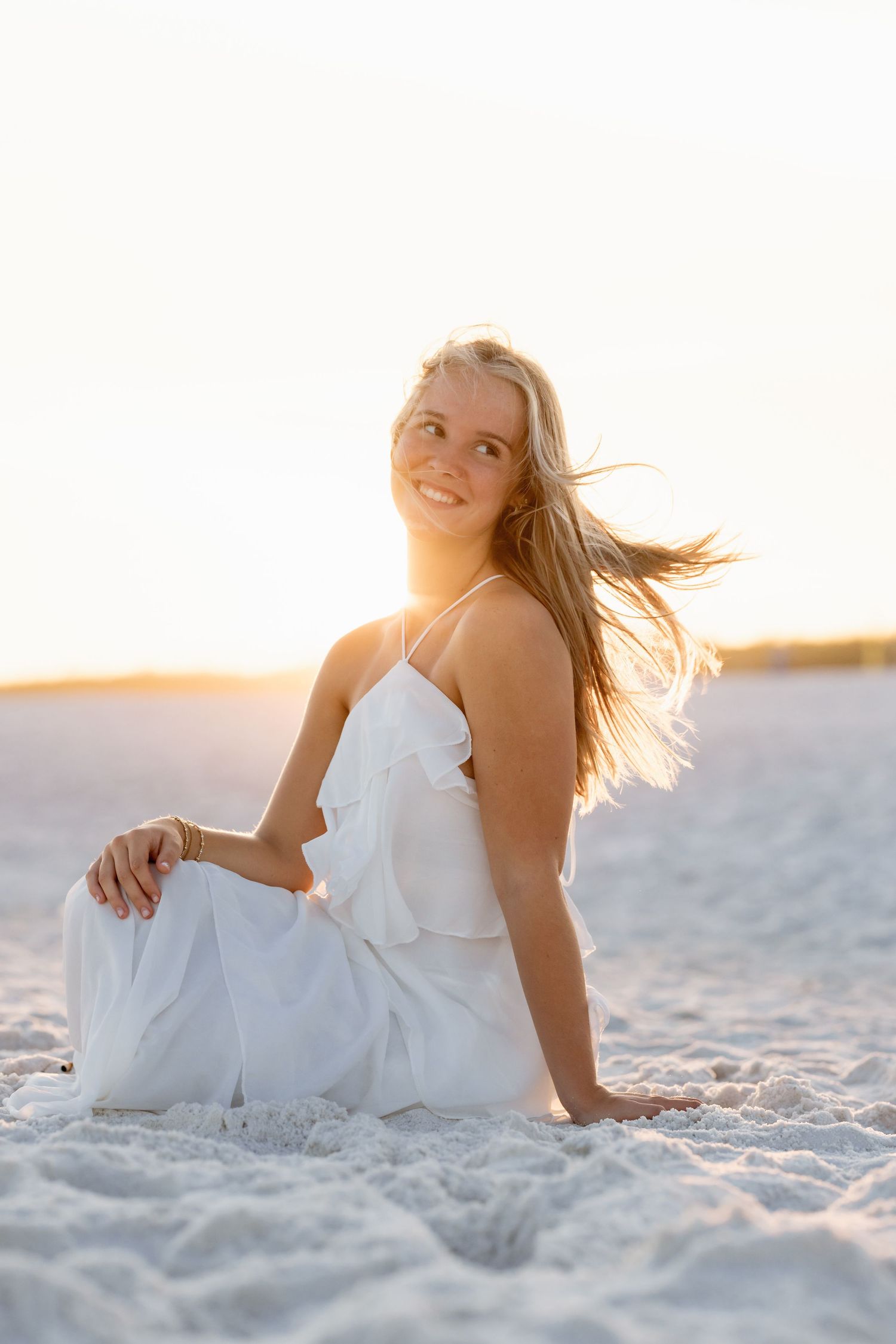 Blonde woman in white dress sitting on sandy beach at golden sunset.