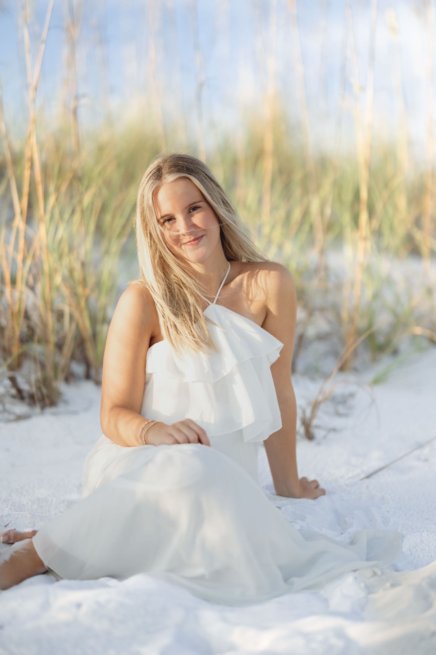 Young blonde woman in white ruffle dress sitting on white sand beach dunes, smiling softly at camera.