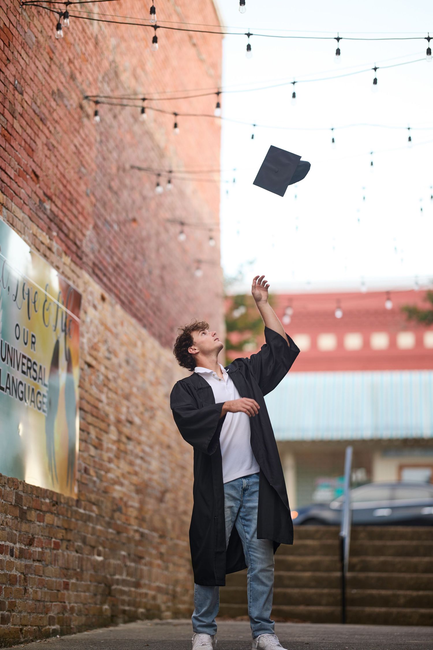 Graduate in black gown tosses cap in air outdoors near brick building with string lights above.