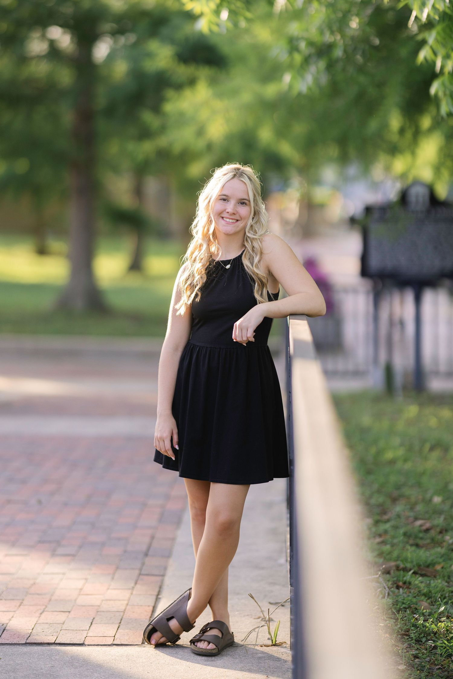 Young woman in black dress leaning on railing in park, smiling with long blonde hair.