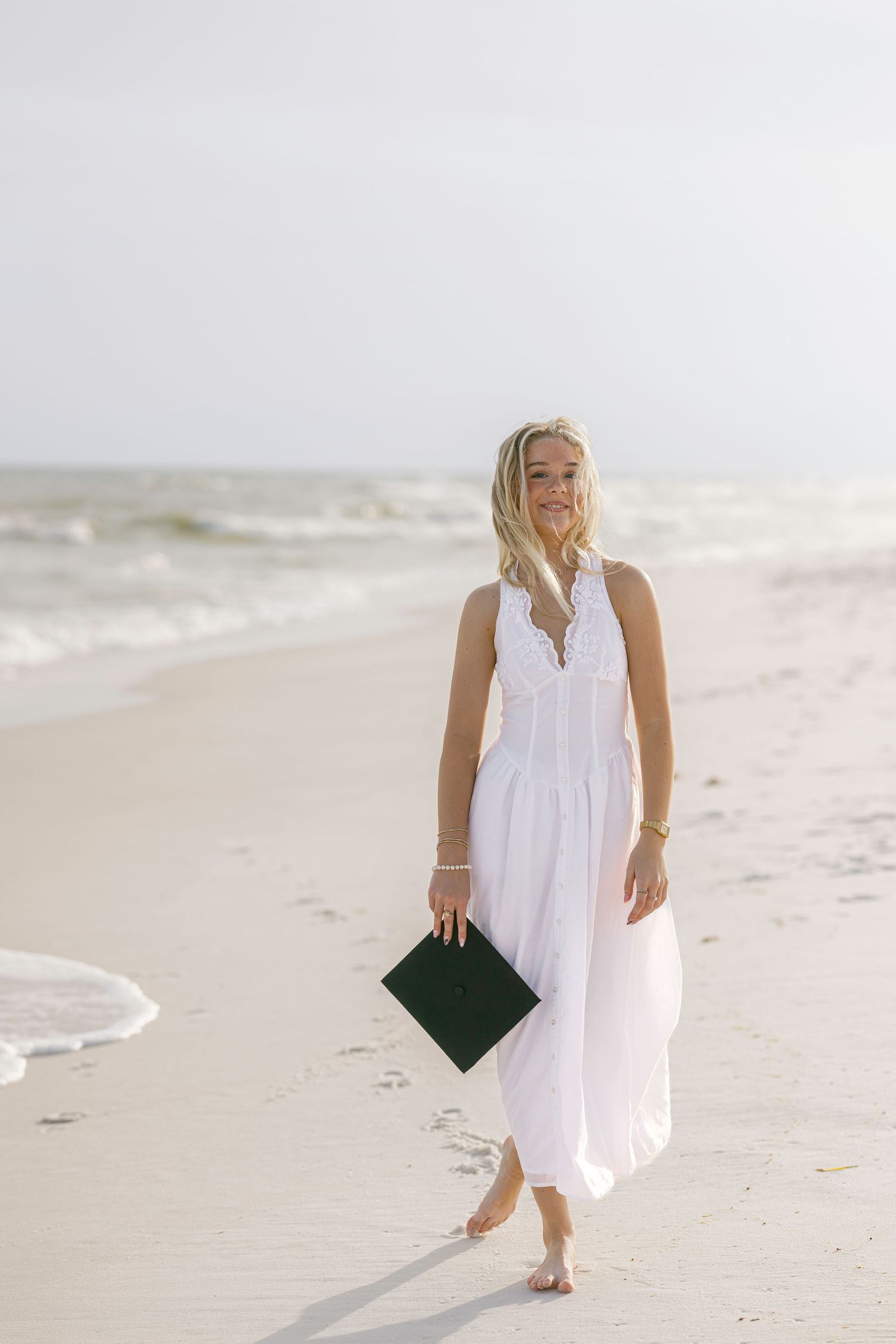 Elegant beach walk scene with white maxi dress and black clutch against ocean waves.
