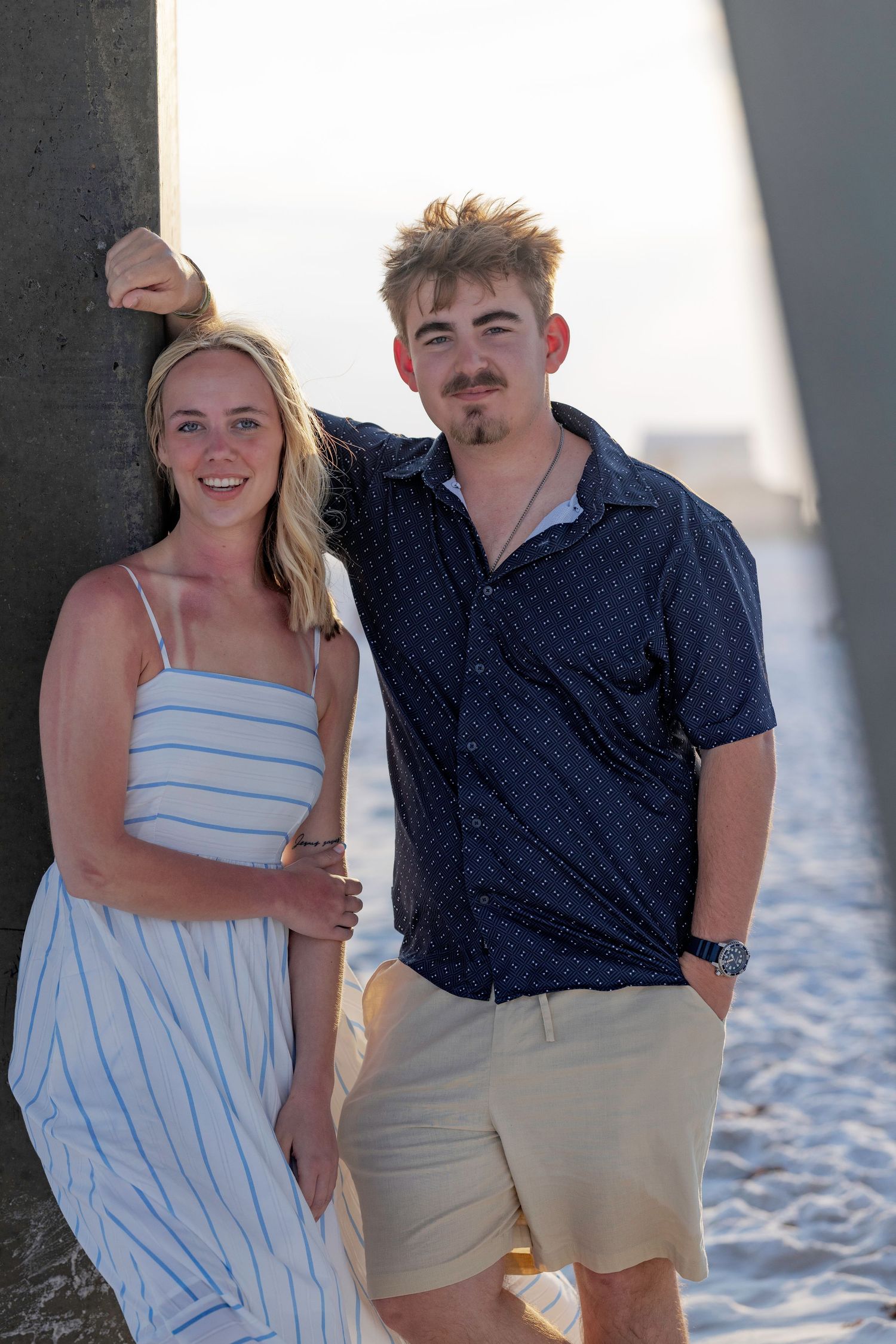 A smiling young couple poses together near a beach pier, woman in striped dress, man in navy shirt and khaki shorts.