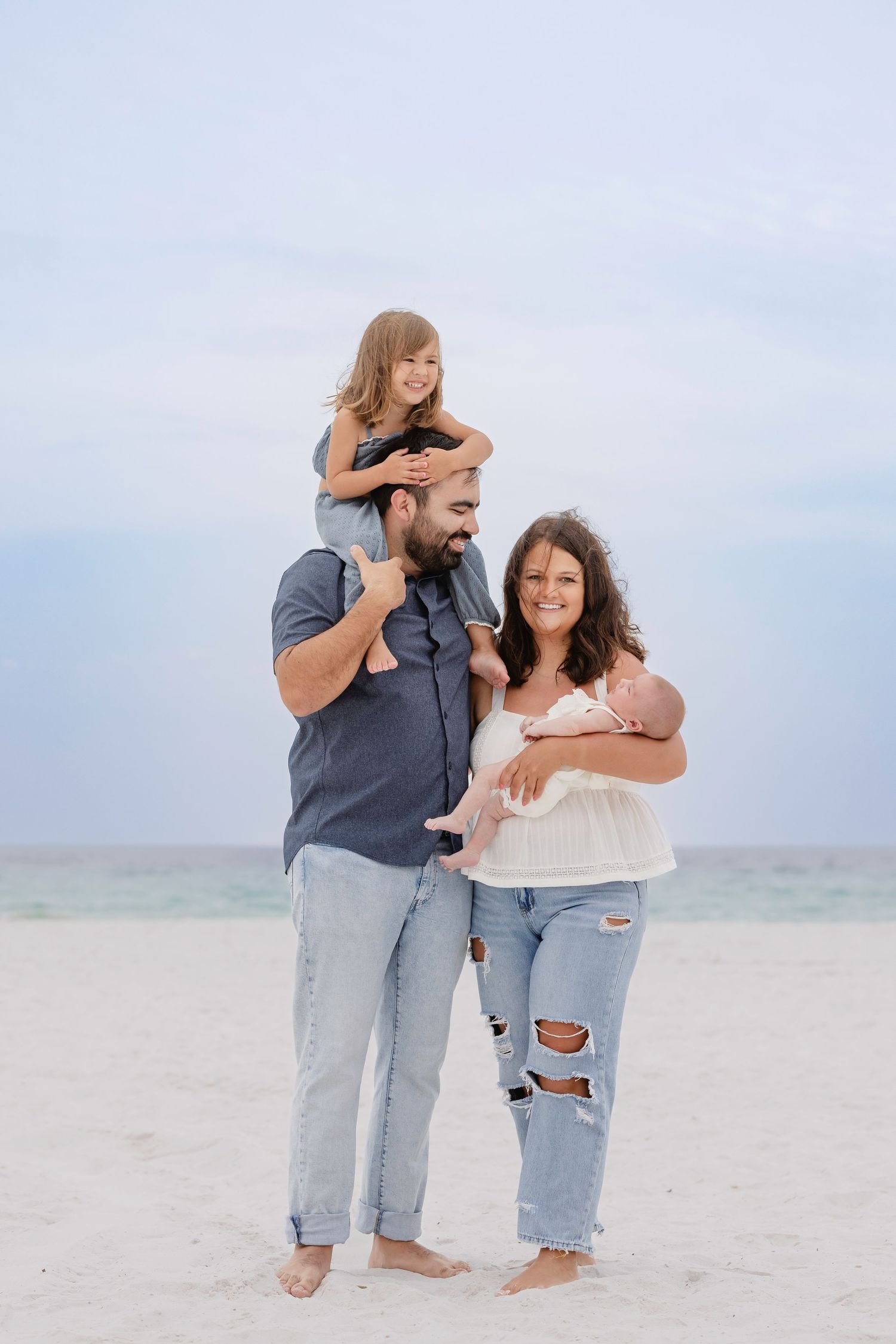 Family of four poses on a sandy beach, dad holds toddler on shoulders while mom cradles newborn baby.