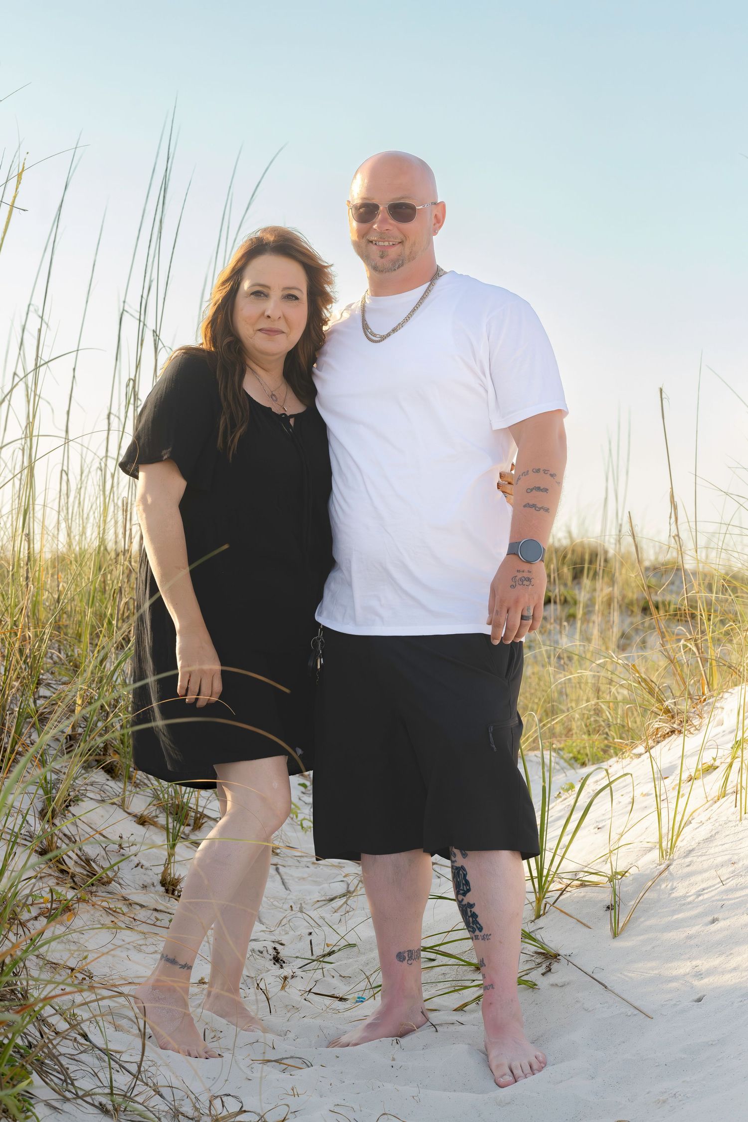 Couple posing together on sandy beach dunes at sunset, man in white shirt and woman in black dress.