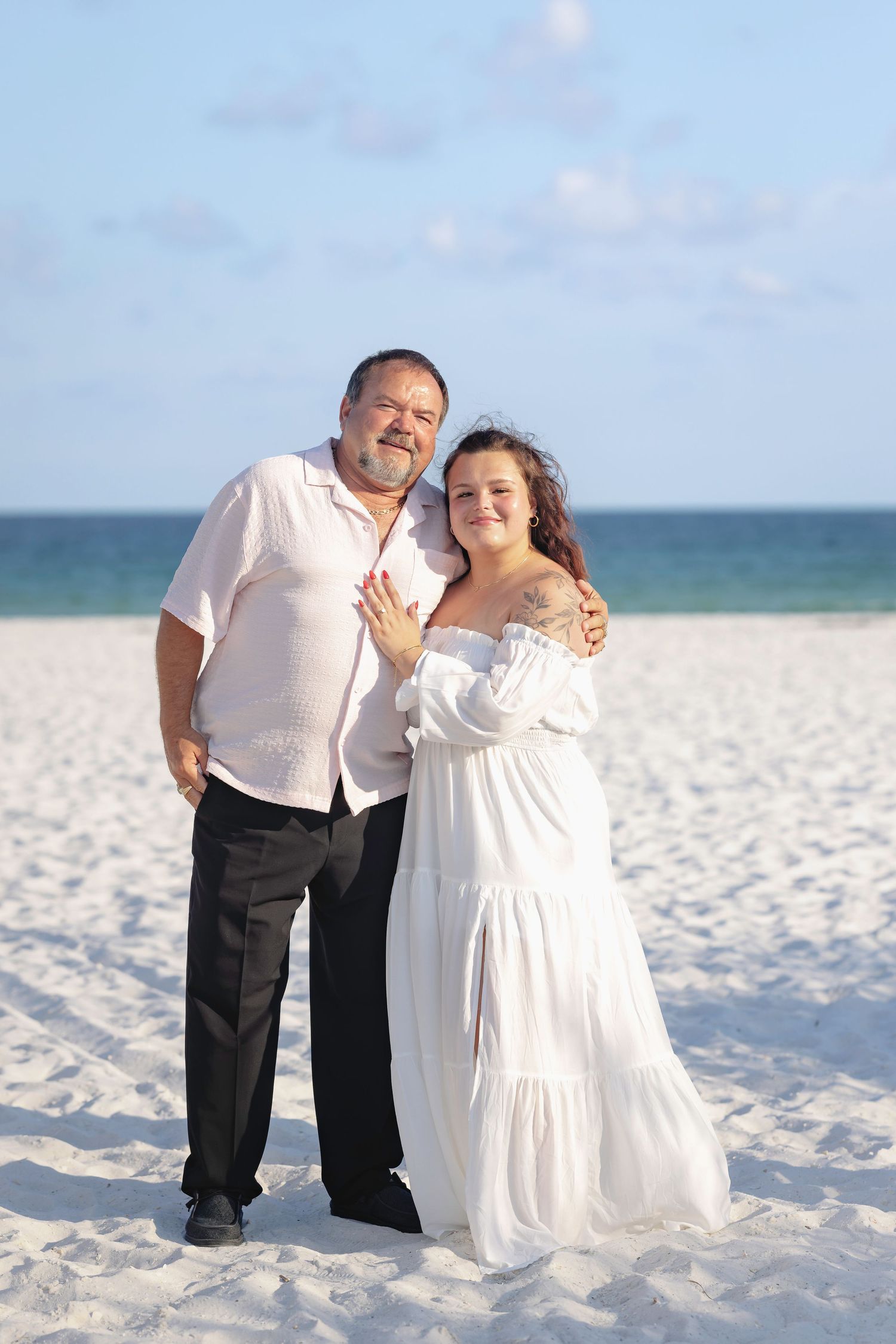 Couple posing on white sand beach, woman in white off-shoulder dress showing ring, man in white shirt and black pants.