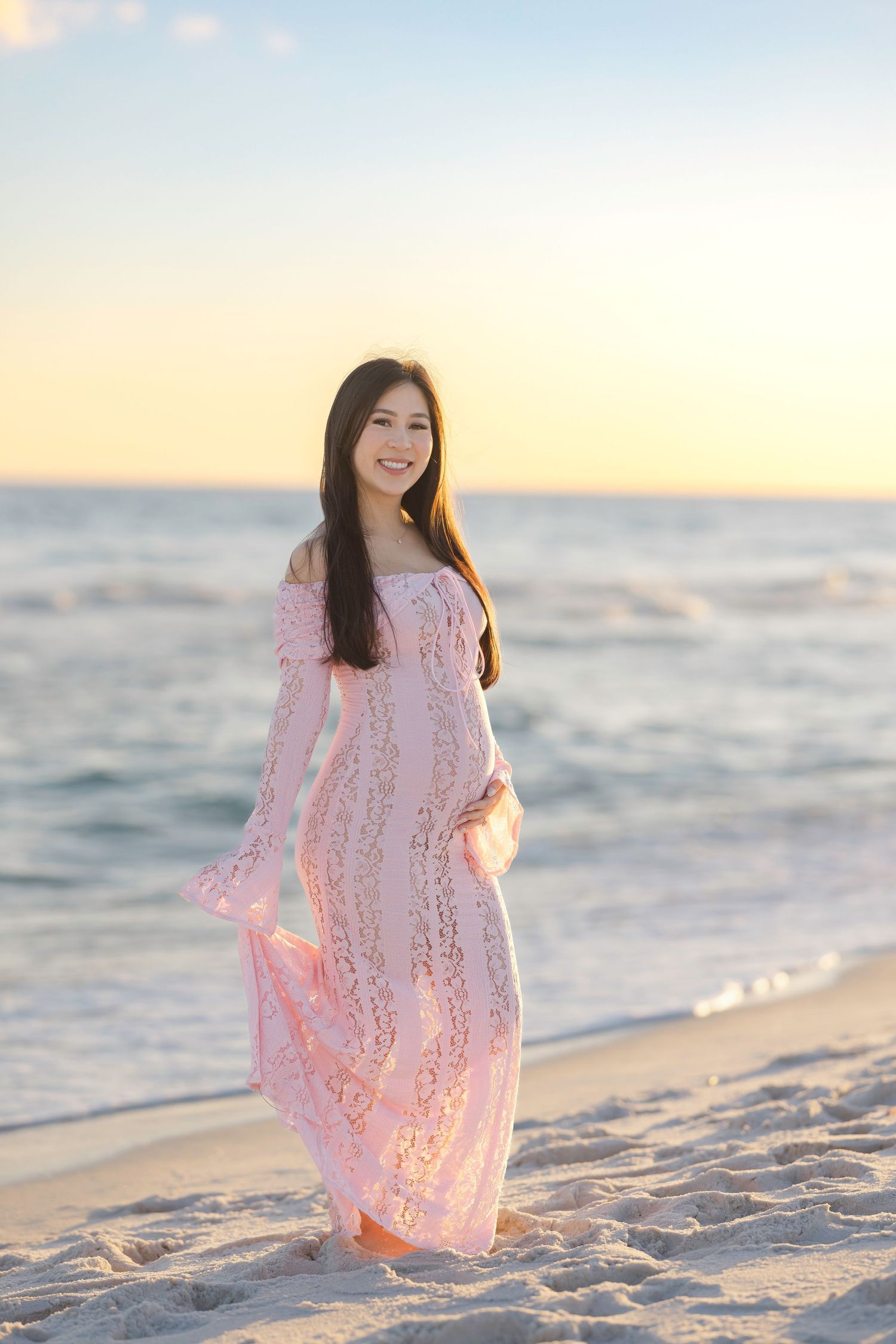 Beautiful woman in pink lace off-shoulder dress posing on sandy beach at golden sunset.