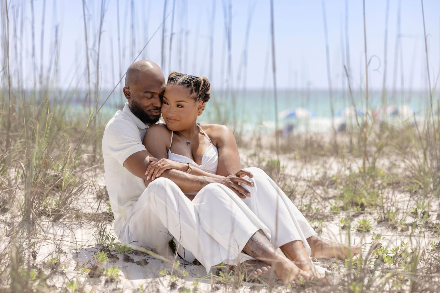 A romantic couple sits together among beach dunes, dressed in white, embracing tenderly.