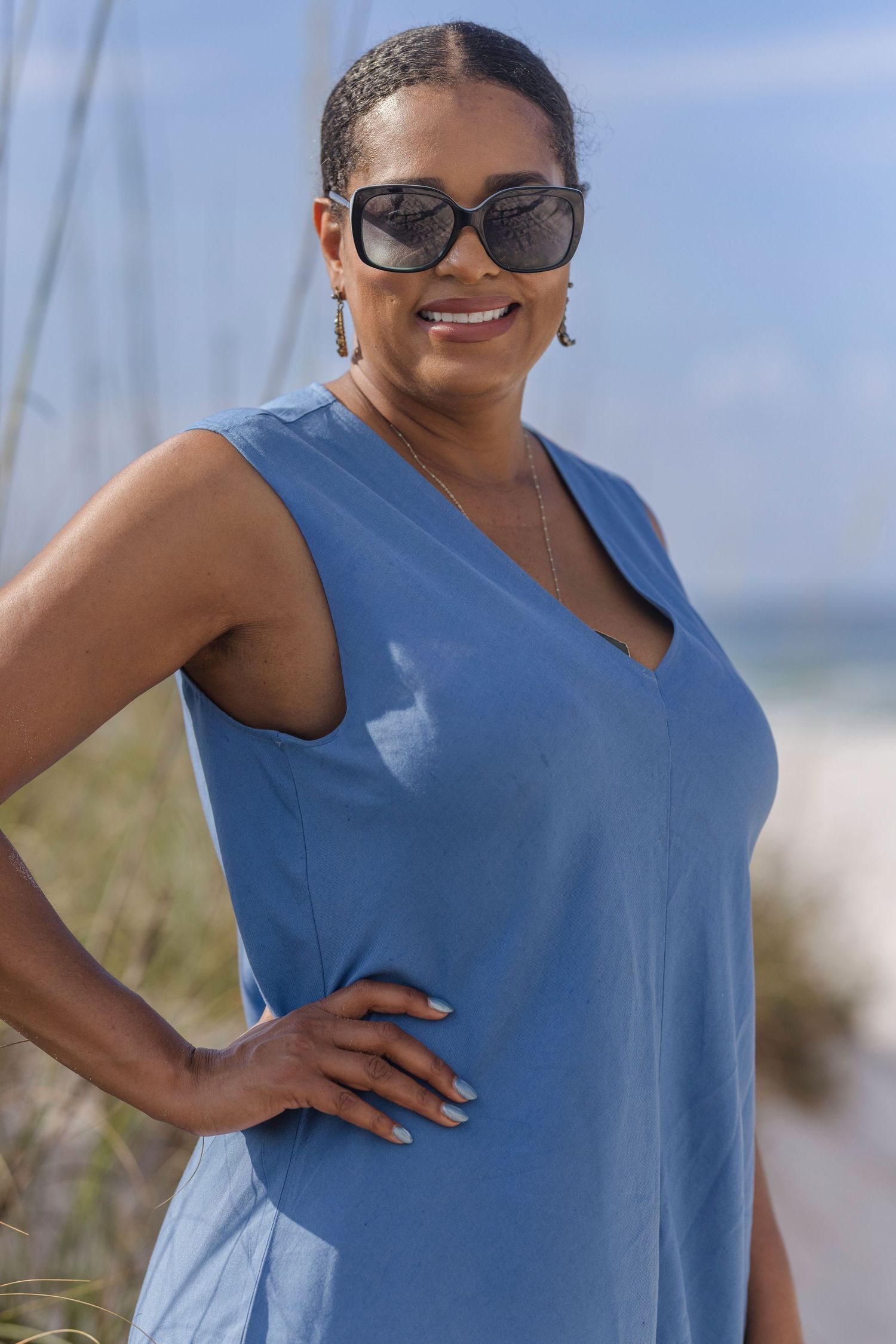 A smiling Black woman in a blue sleeveless dress and sunglasses posing on a sunny beach.