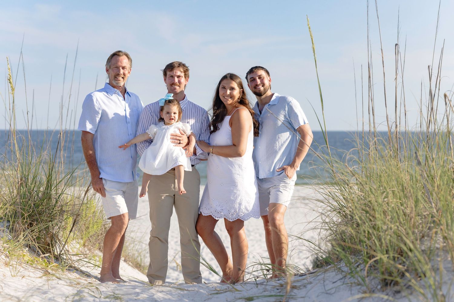 Family of five posing together on a sandy beach path surrounded by sea grass on a sunny day.